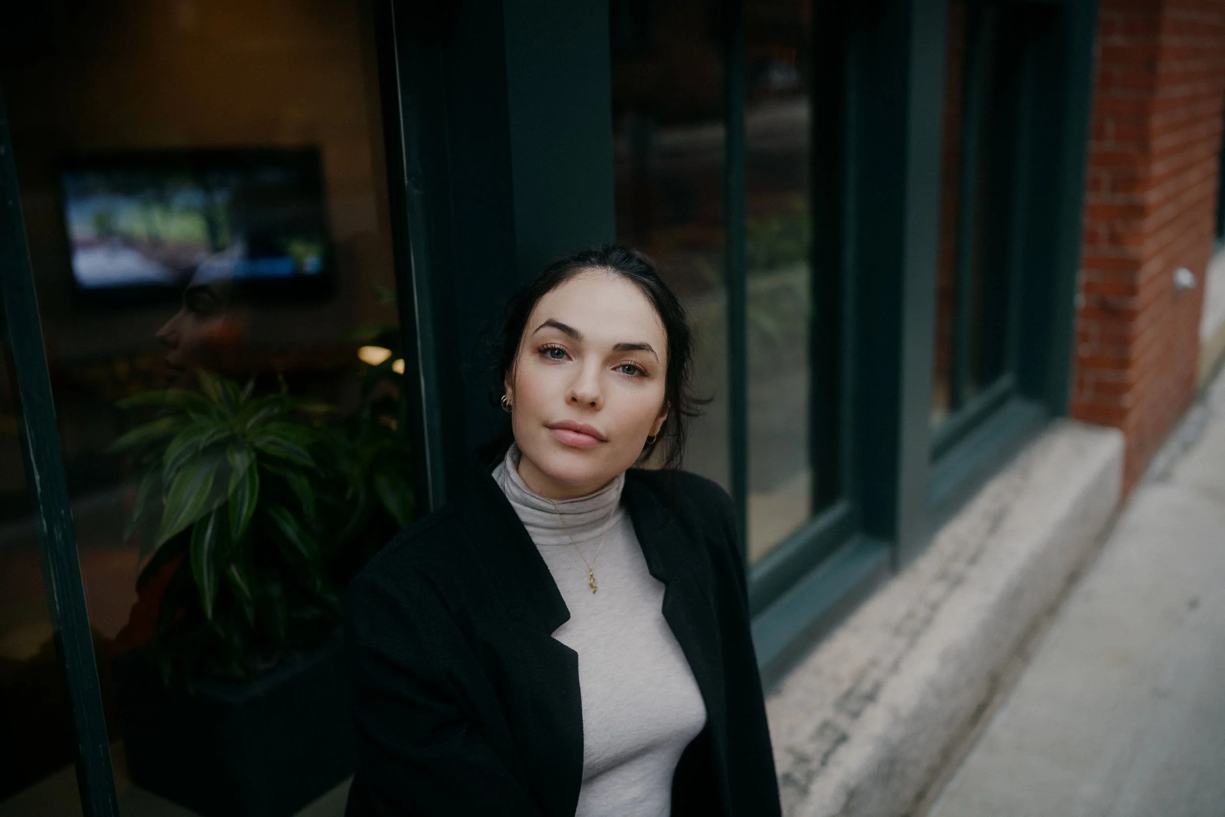 A woman with dark hair and fair skin leaning against a glass window, wearing a light turtleneck and a black blazer, looking at the camera.