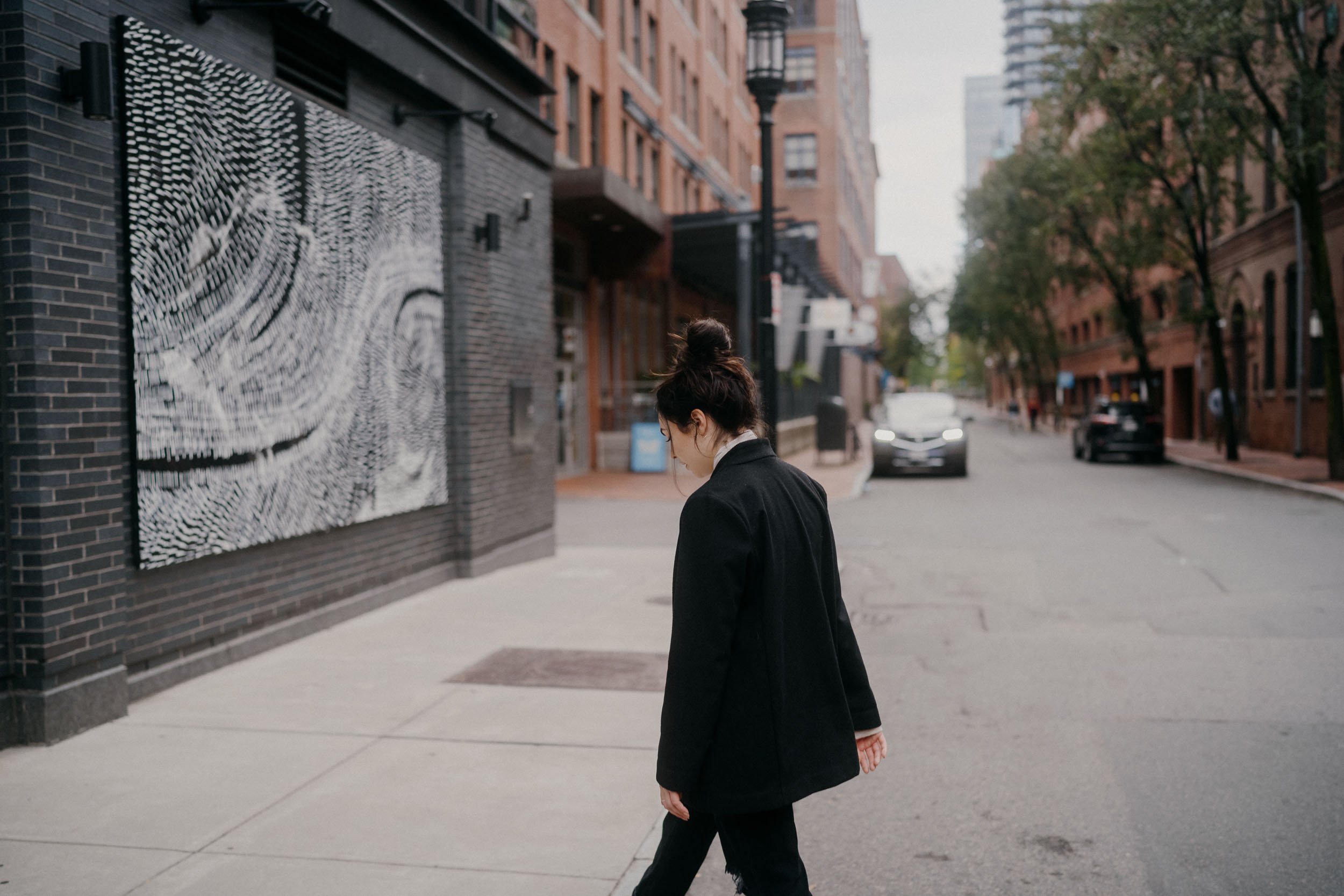 A woman in a black coat walking on a city sidewalk next to a building with a large black and white abstract art piece. Trees line the street and cars are visible in the background.