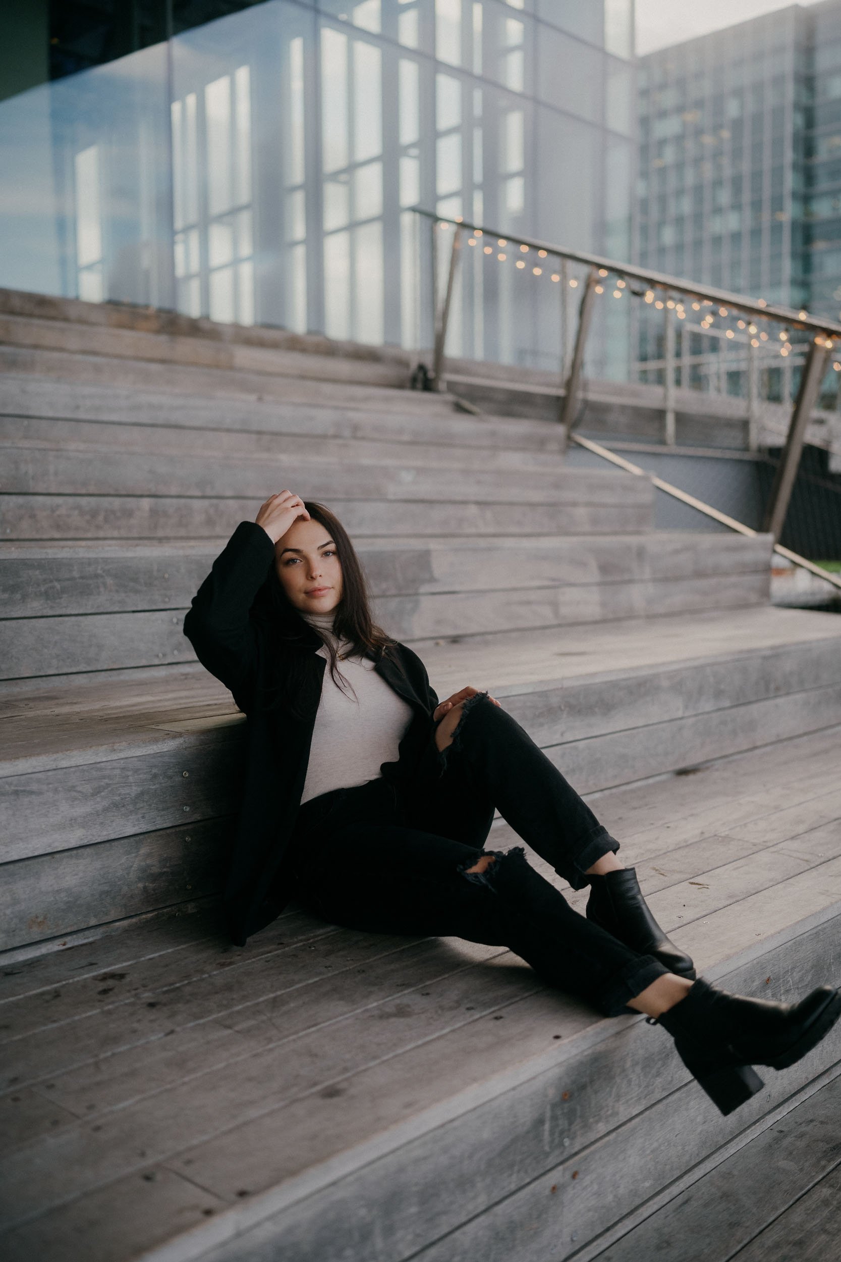 Young woman with dark hair dressed in a black jacket, white top, black ripped jeans, and black boots, sitting on wooden steps outdoors, with modern glass buildings and string lights in the background.