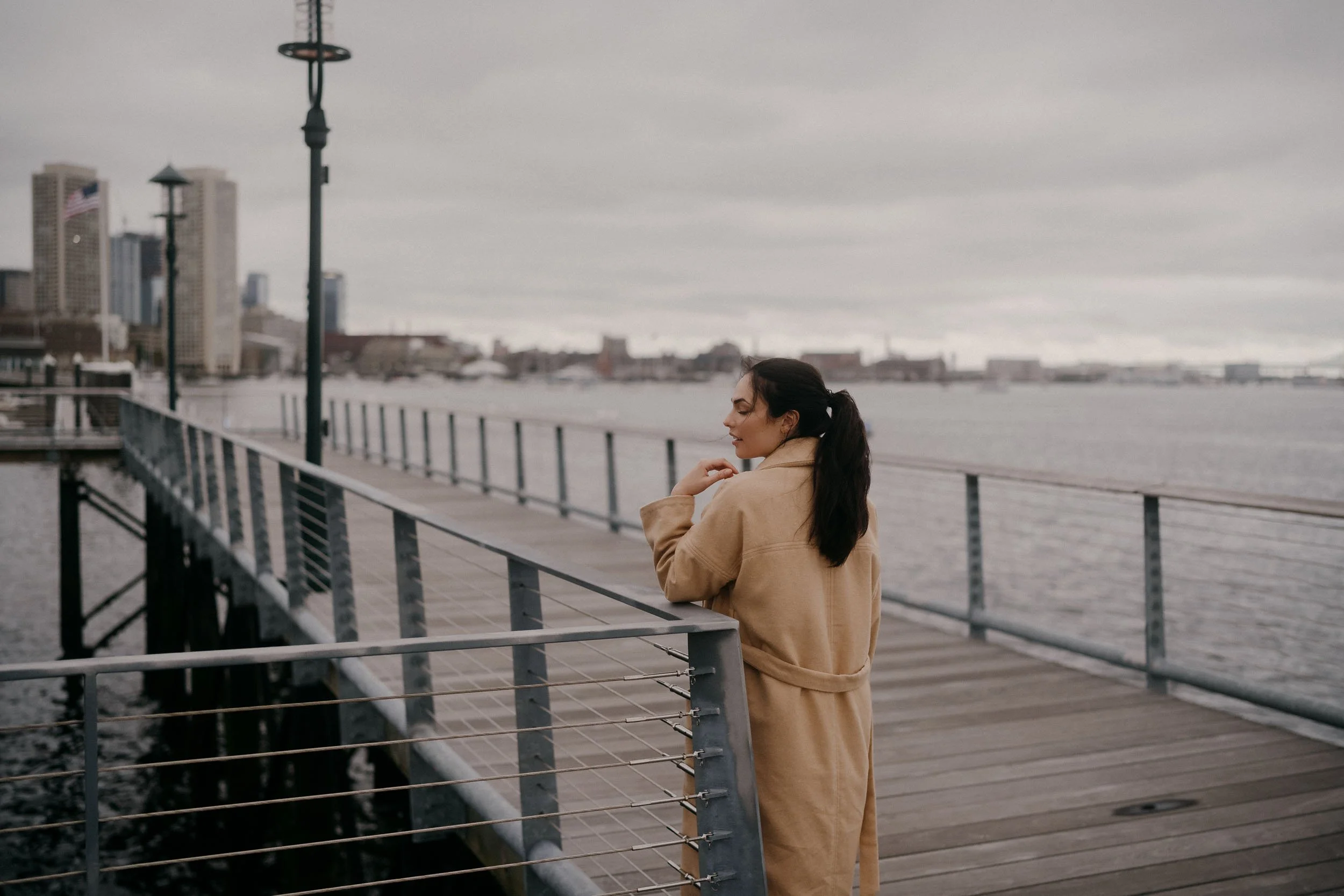 A woman in a beige coat standing on a wooden dock by the water, with a city skyline in the background under a cloudy sky.