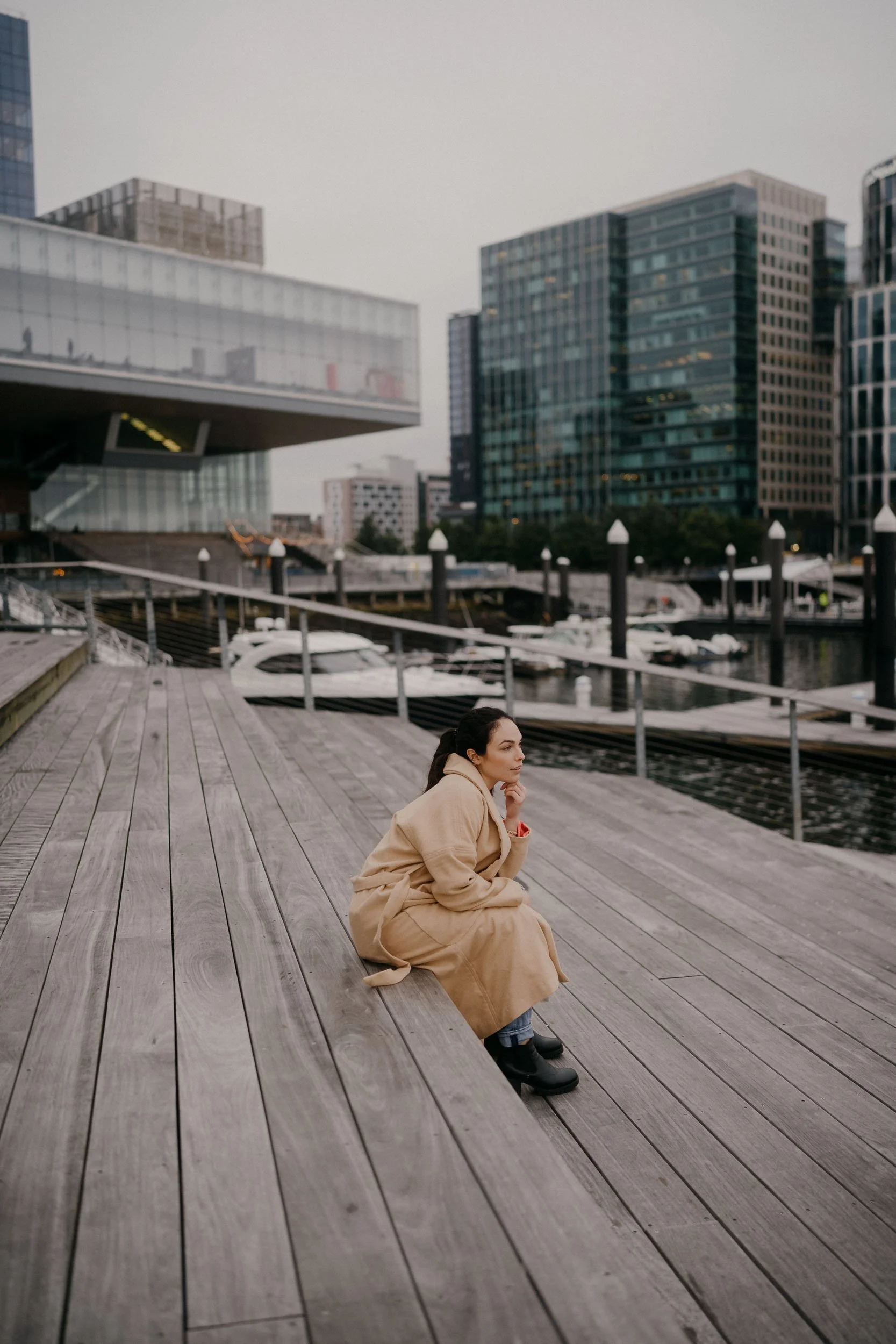 A woman in a beige coat and black boots sitting on a wooden dock by the water, with modern buildings and boats in the background.