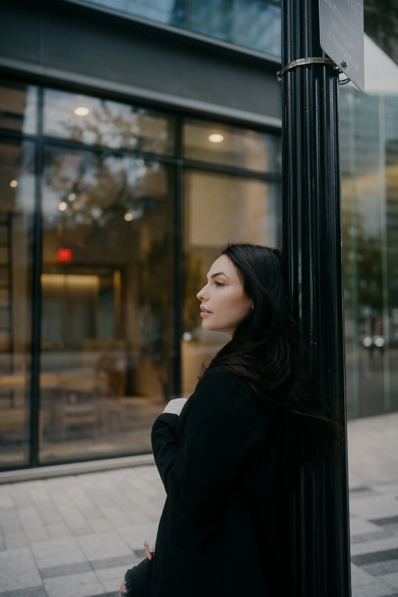 A woman with long dark hair leaning against a black pole outside a modern building with glass windows.