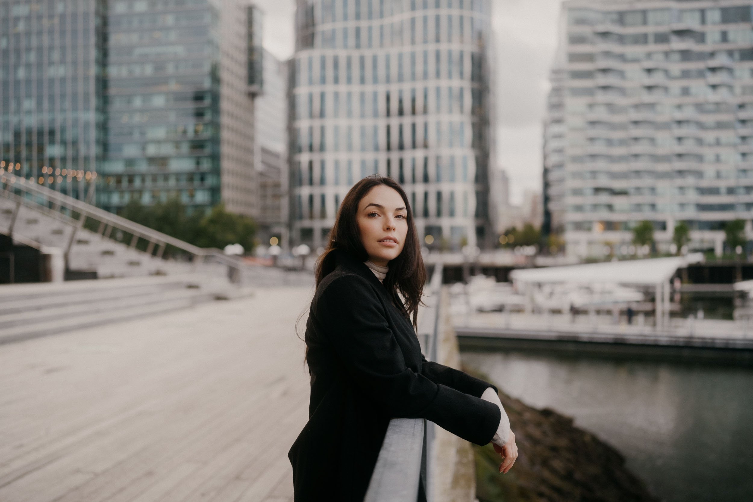 A woman with dark hair leaning on a railing along a city waterfront with modern high-rise buildings in the background.