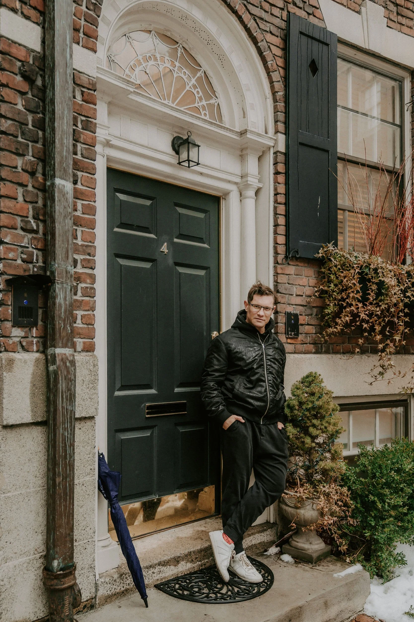 A man wearing a black jacket and pants leaning against a black front door of a brick house.