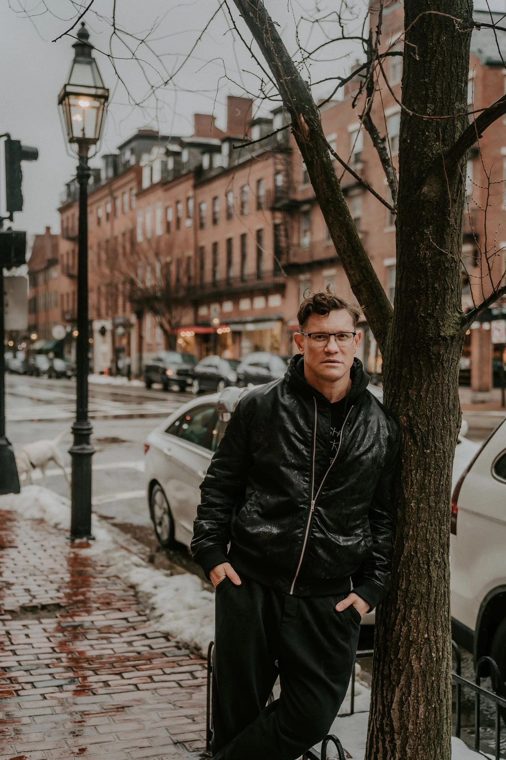 A man in a black jacket and glasses standing next to a tree on a city sidewalk on a rainy day.