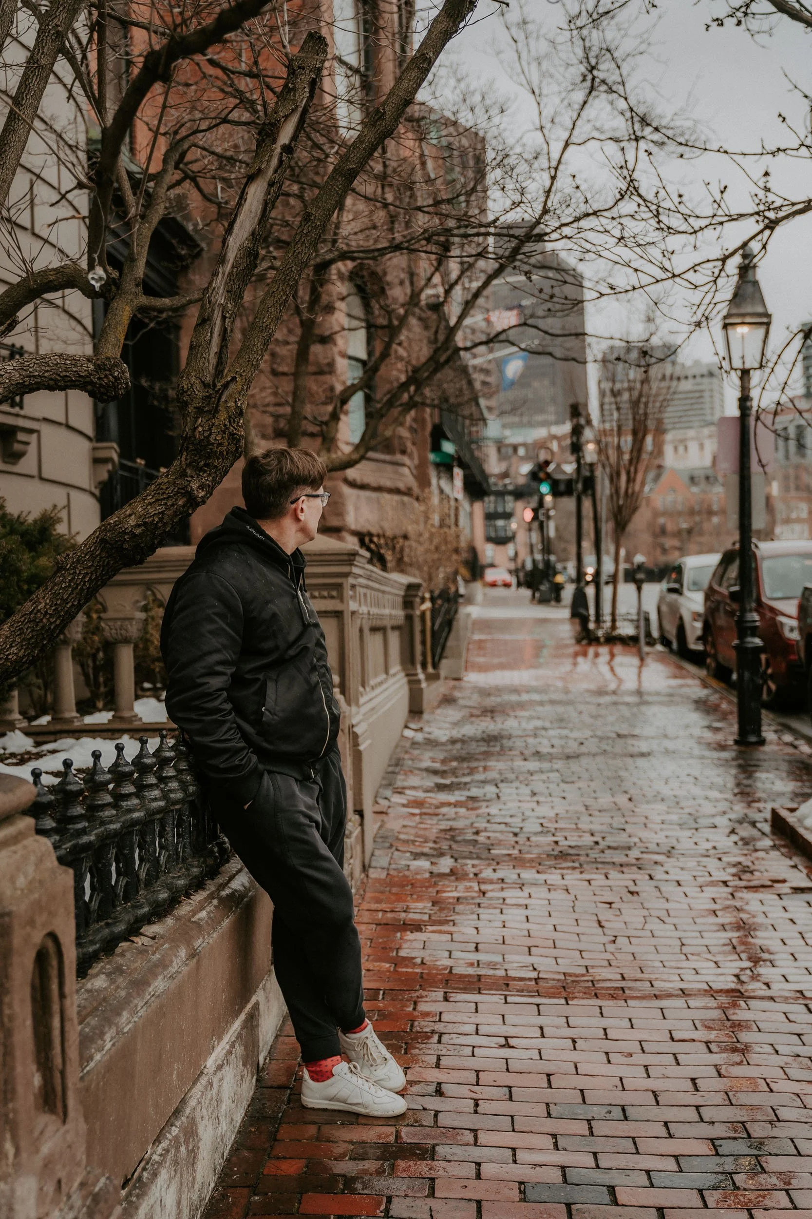 A man in a black jacket and white sneakers leans against a railing on a brick sidewalk in an urban area with bare trees and parked cars, during dusk or overcast weather.