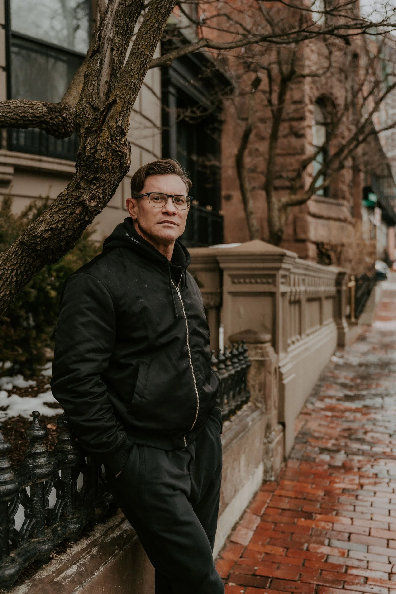 A man wearing glasses and a black jacket standing on a sidewalk near a tree and brownstone buildings.