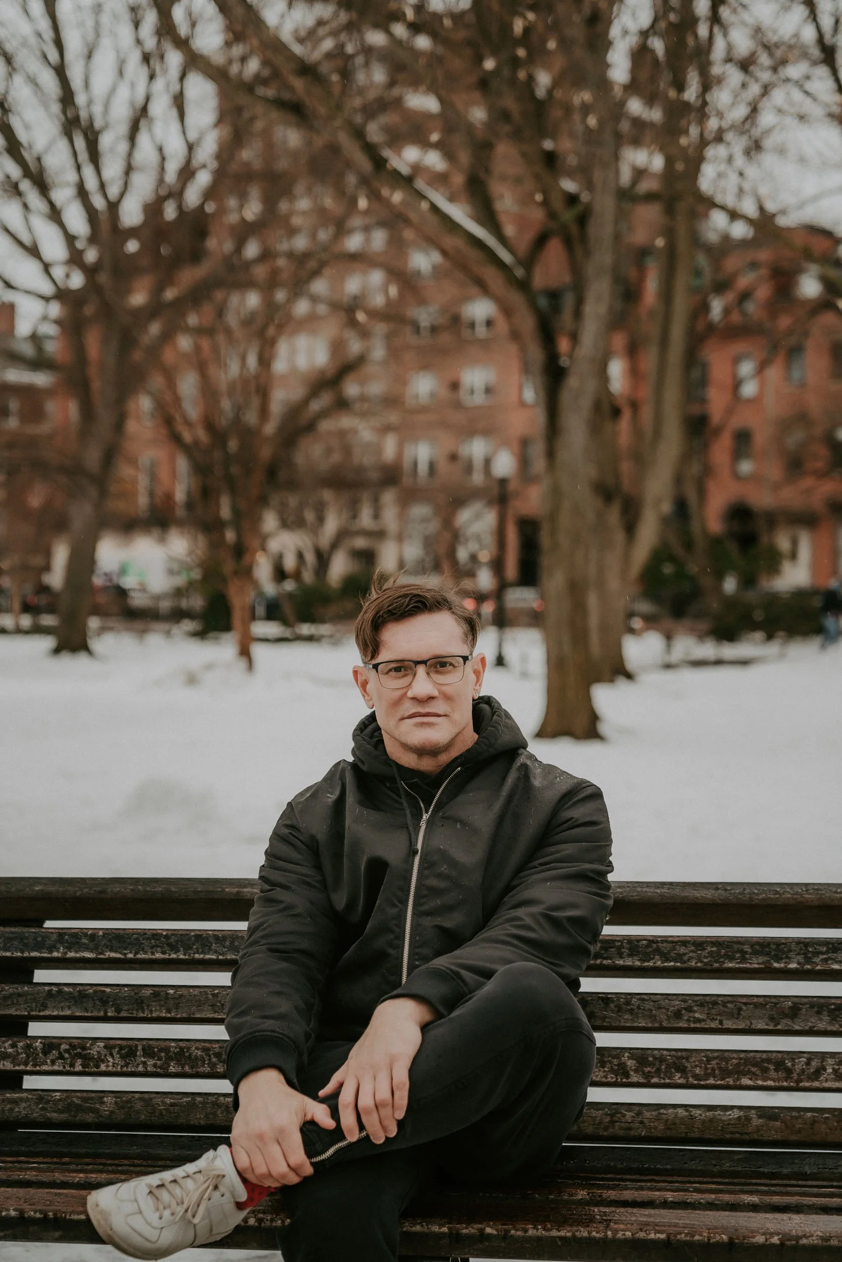 A man sitting on a wooden park bench in winter, wearing glasses, a black jacket, and sneakers, with leafless trees and a red brick apartment building in the background.
