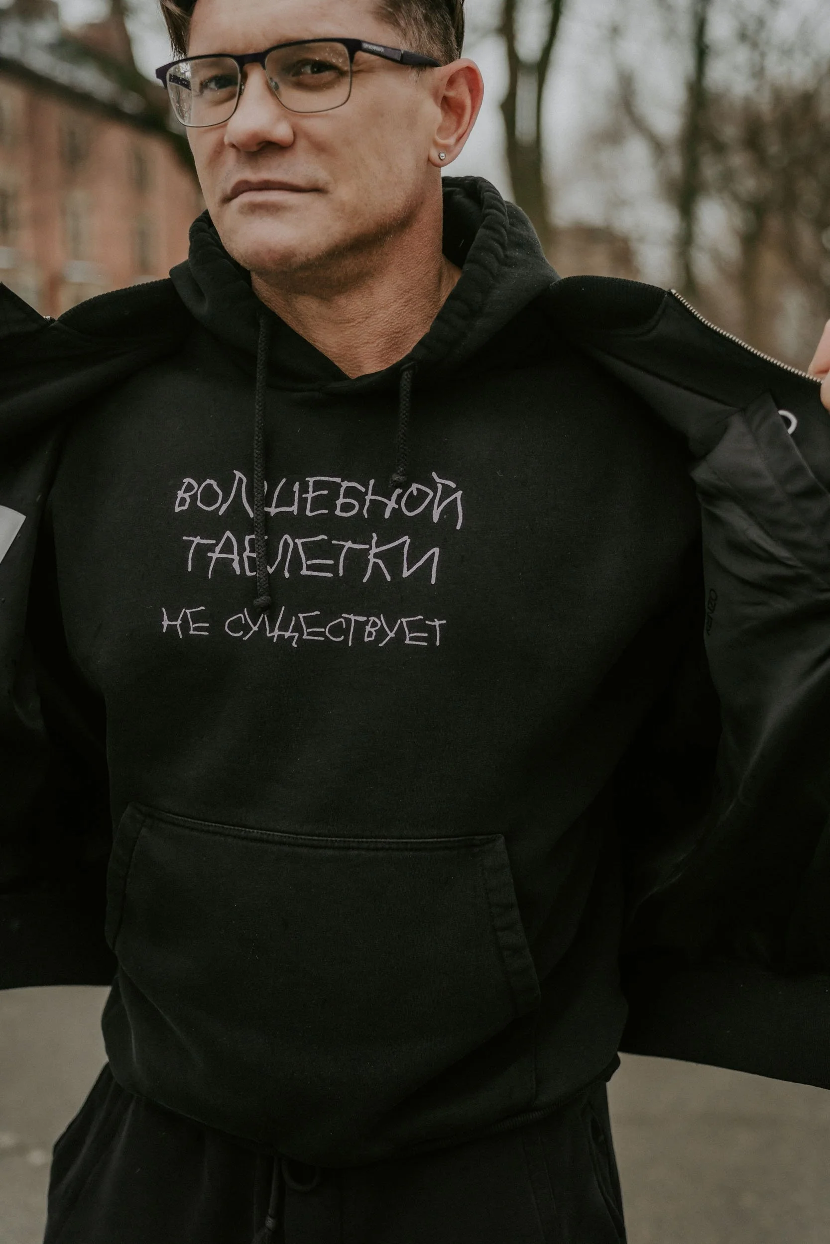 A man wearing glasses and a black hoodie with Russian text on it, standing outdoors, with trees and buildings in the background.