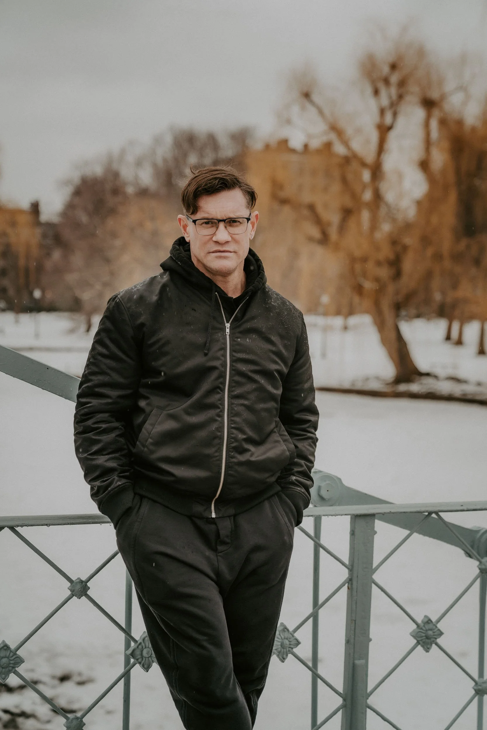 A man in a black jacket and pants standing outdoors on a bridge with snow and bare trees in the background.