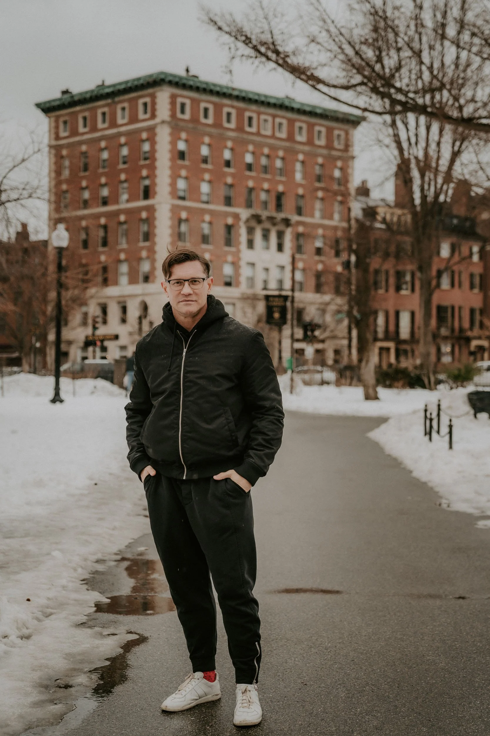 A man in a black jacket and sweatpants standing outdoors on a snowy pathway in an urban setting, with historic brick buildings and leafless trees in the background.