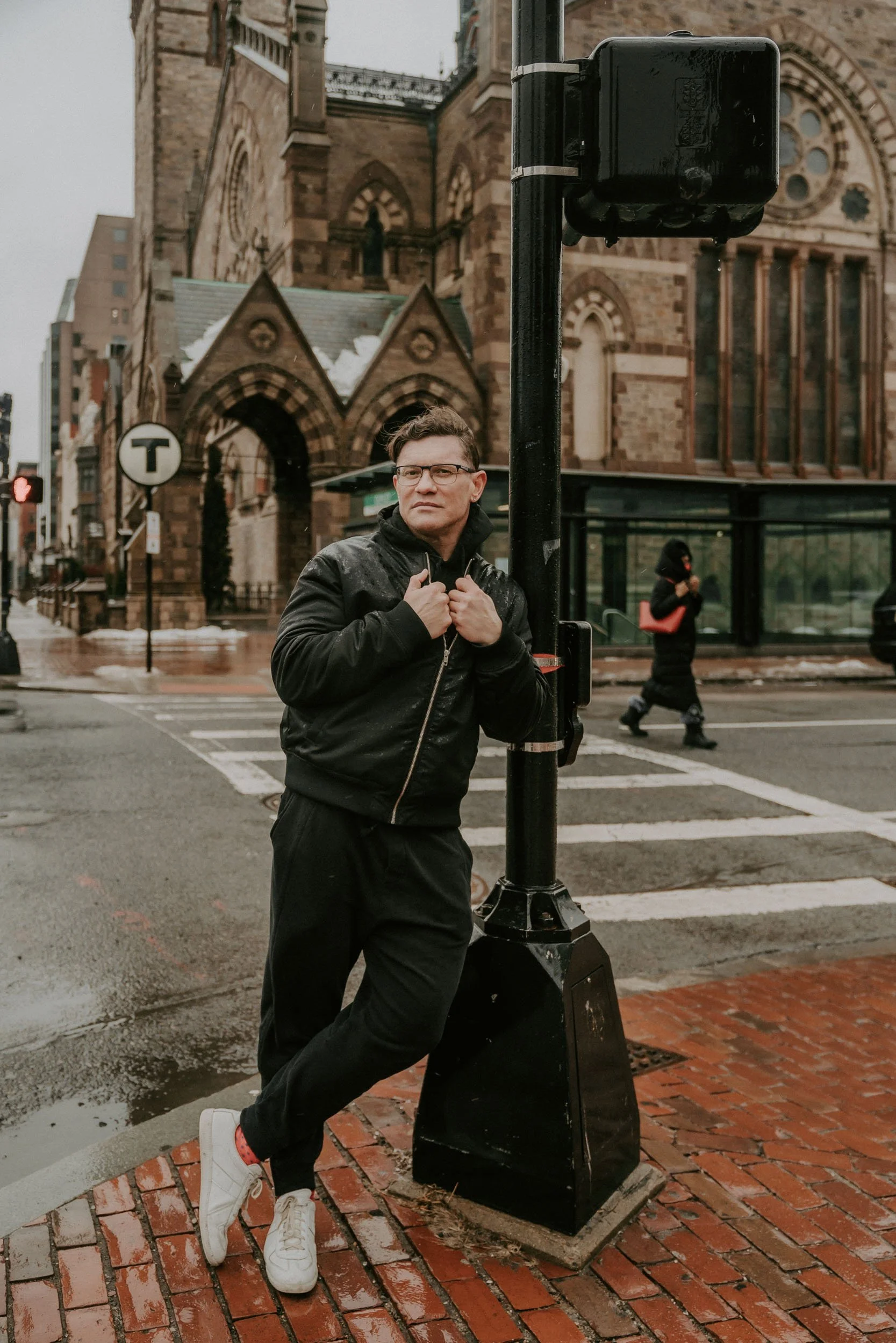 A young man with glasses wearing a black jacket and black pants standing on a brick crosswalk corner on a rainy day in front of an old church.”}