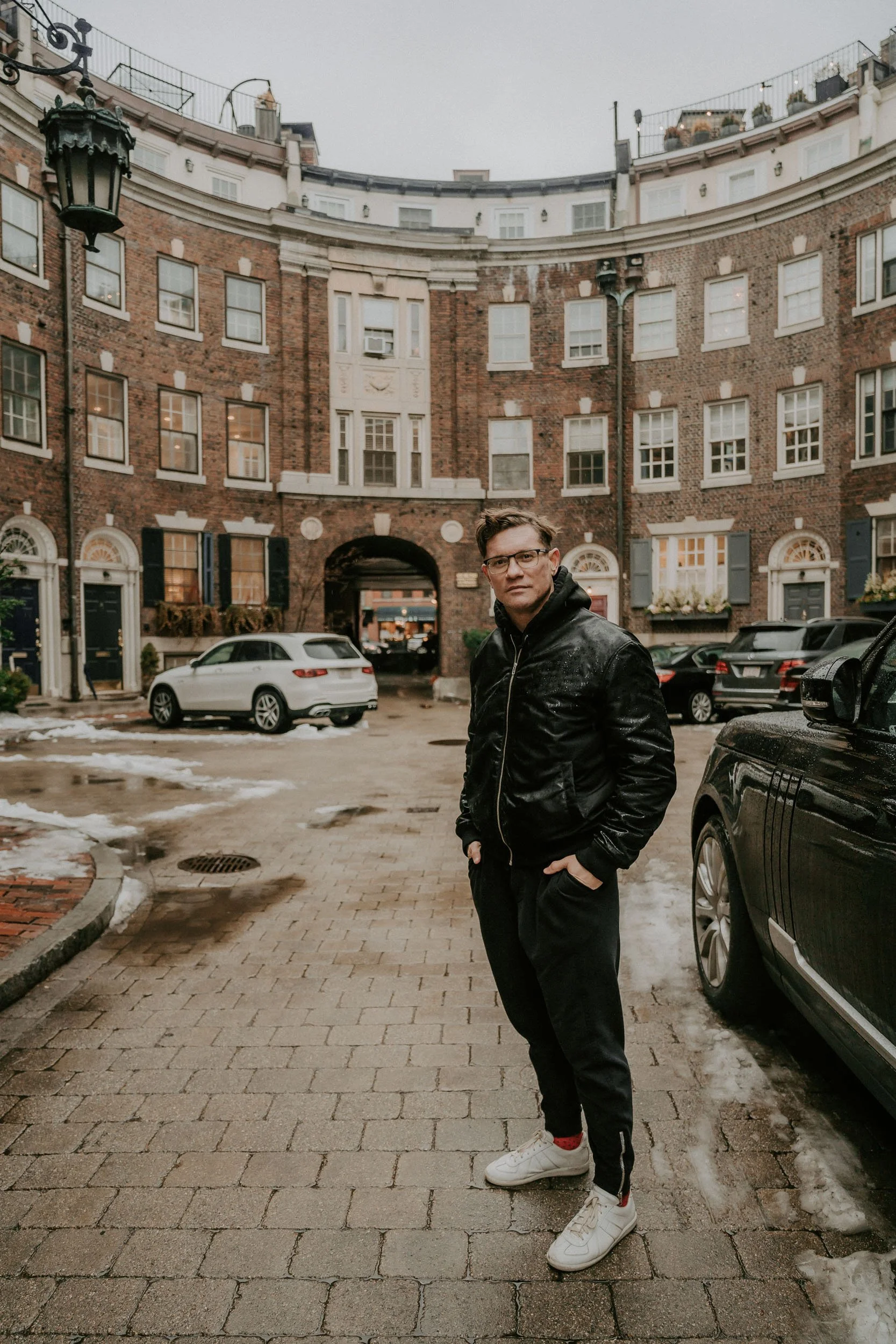 A man wearing glasses, a black jacket, and black pants stands with hands in pockets on a brick-paved street in front of a large, curved brick building with multiple windows. There are parked cars, some snow patches, and a lantern-style street light.