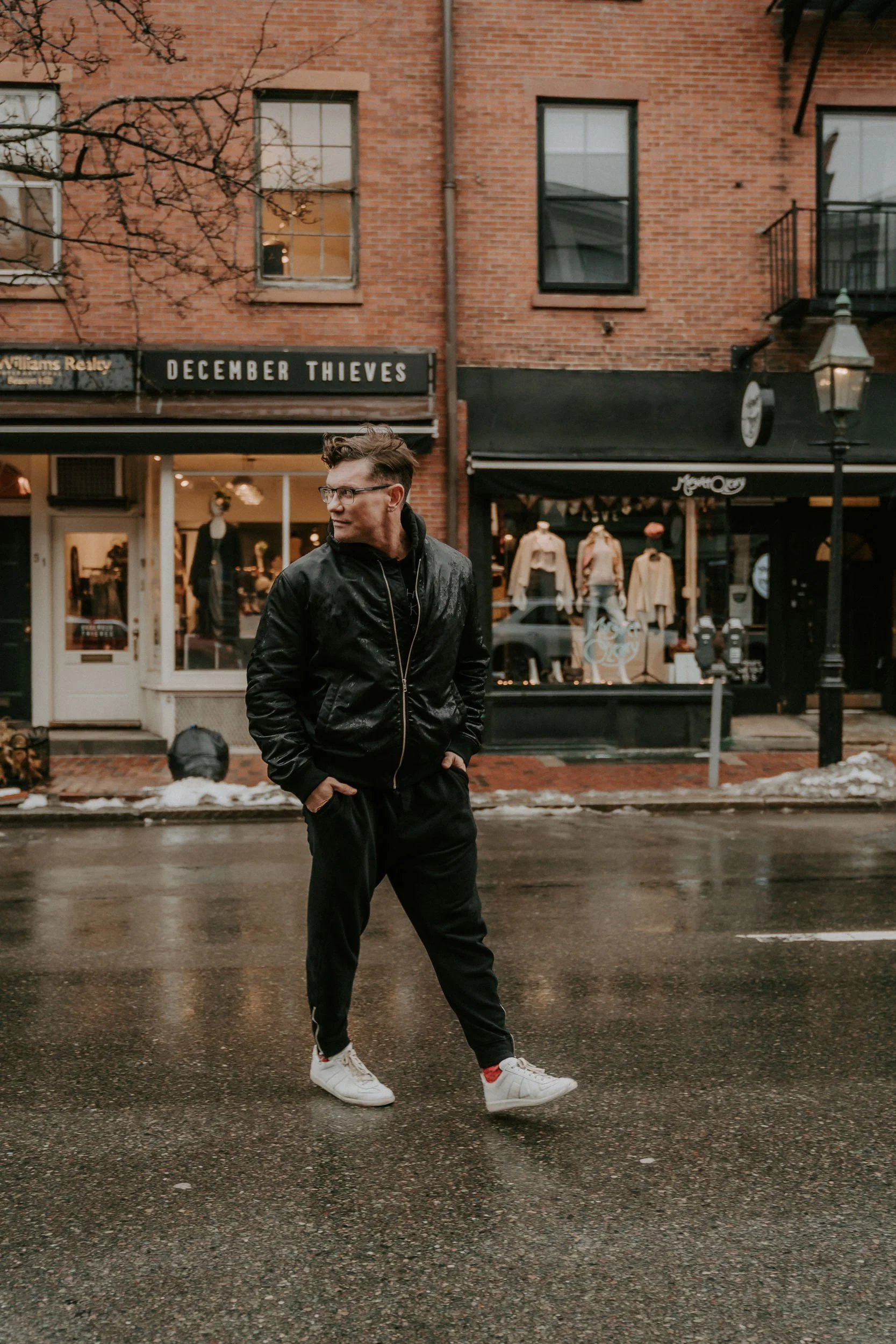 A man walking on a wet city street in front of a store with a sign that reads 'December Thieves', wearing a black jacket, black pants, white sneakers, and glasses, with a brick building in the background.
