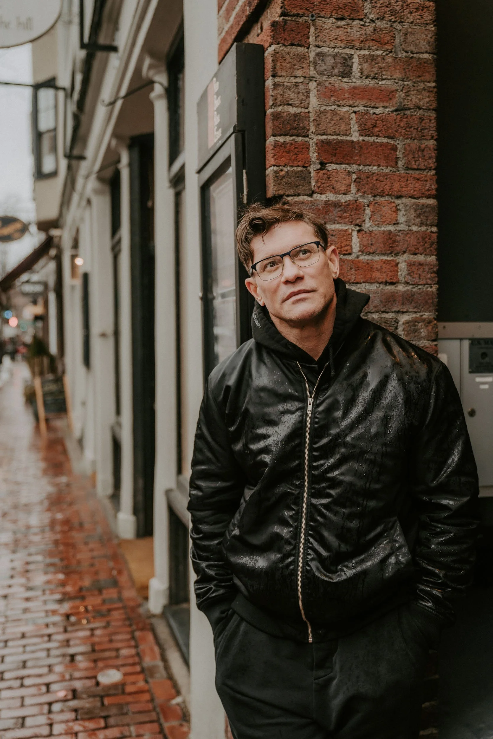 A man with glasses, wearing a black rainy jacket, stands against a brick wall on a rainy street.