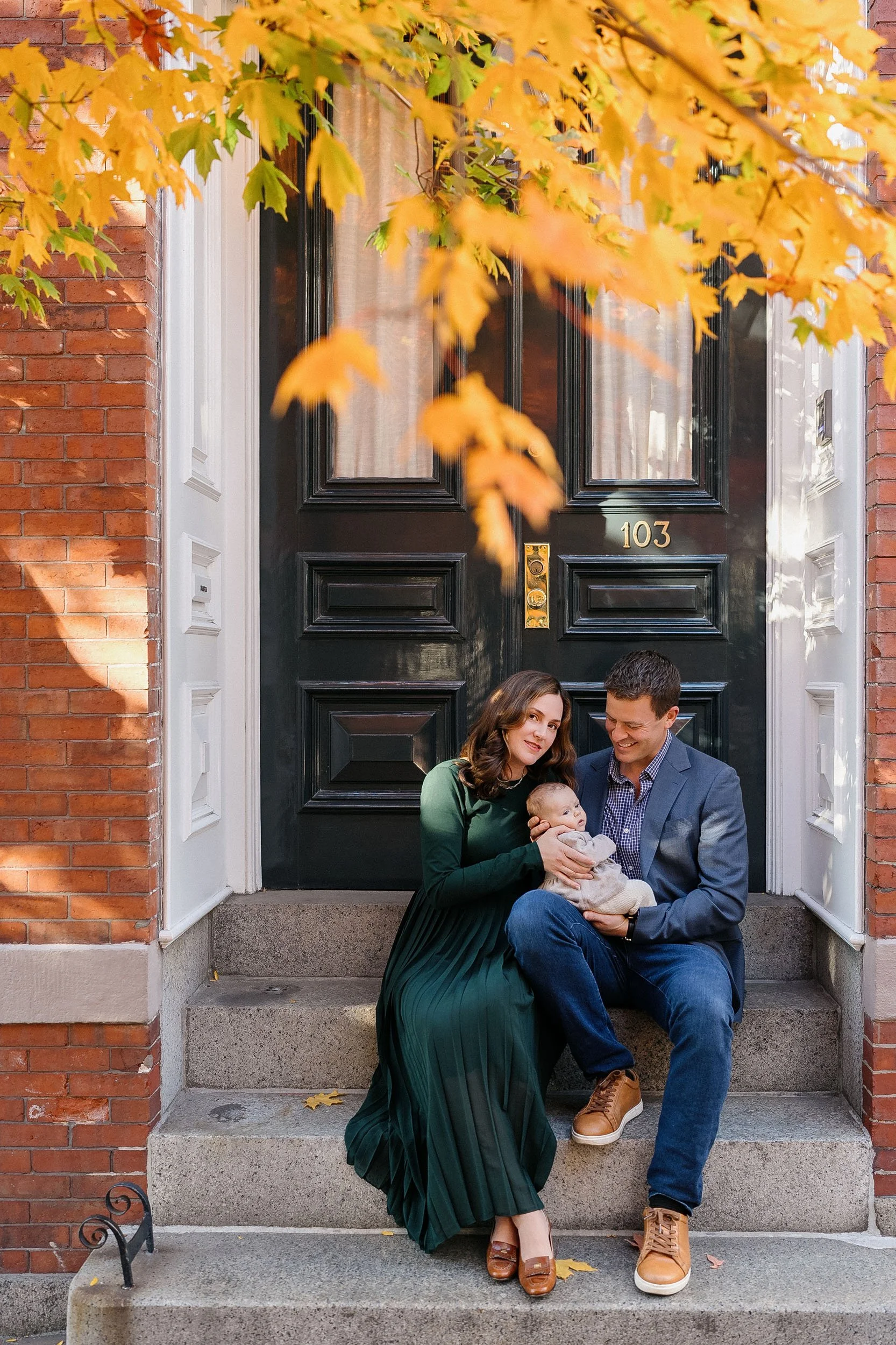 A family of three sitting on the steps of a black front door, with orange and yellow autumn leaves visible above. The woman wears a green dress, the man a gray blazer with jeans, and they hold a baby.