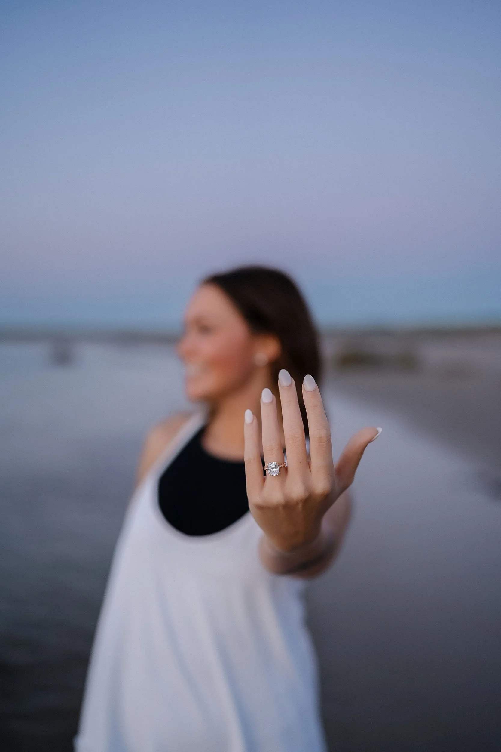 A woman on the beach showing off an engagement ring on her finger, with a blurred background of the ocean and sky during dusk.