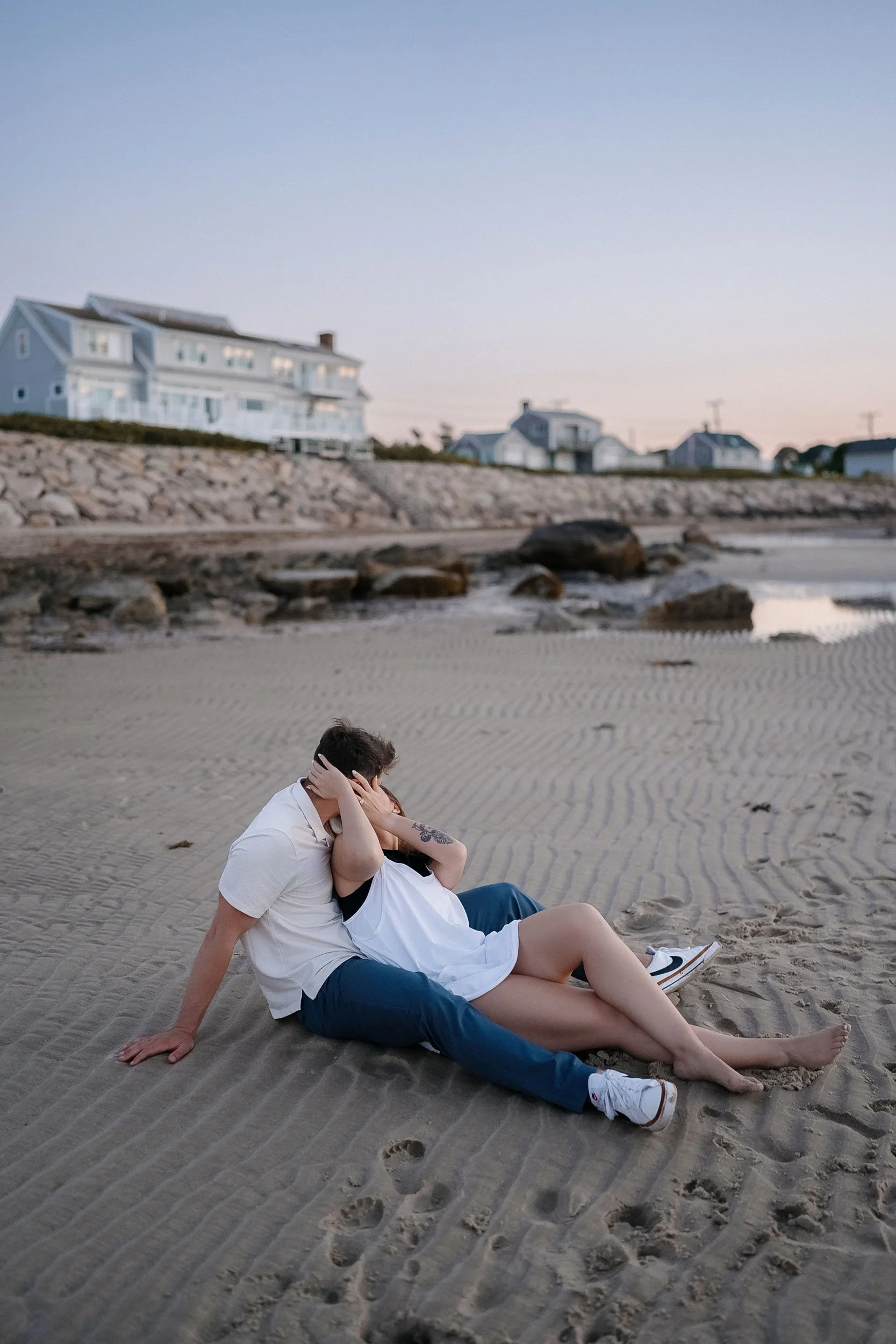 A couple sitting on the sand at the beach, embracing with their faces close together during sunset, with houses and rocks in the background.