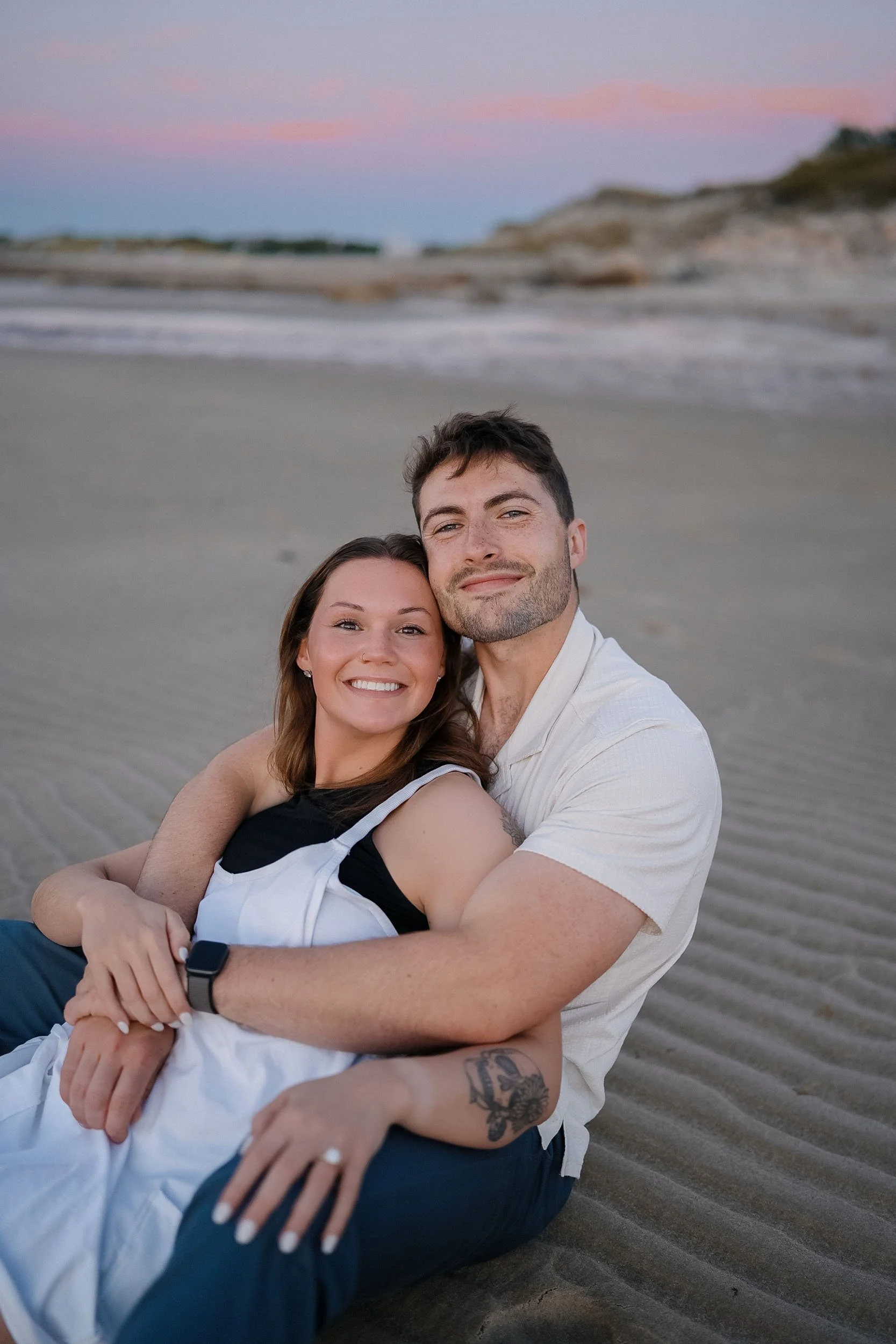 A happy couple sitting on the beach at sunset, smiling and embracing each other.