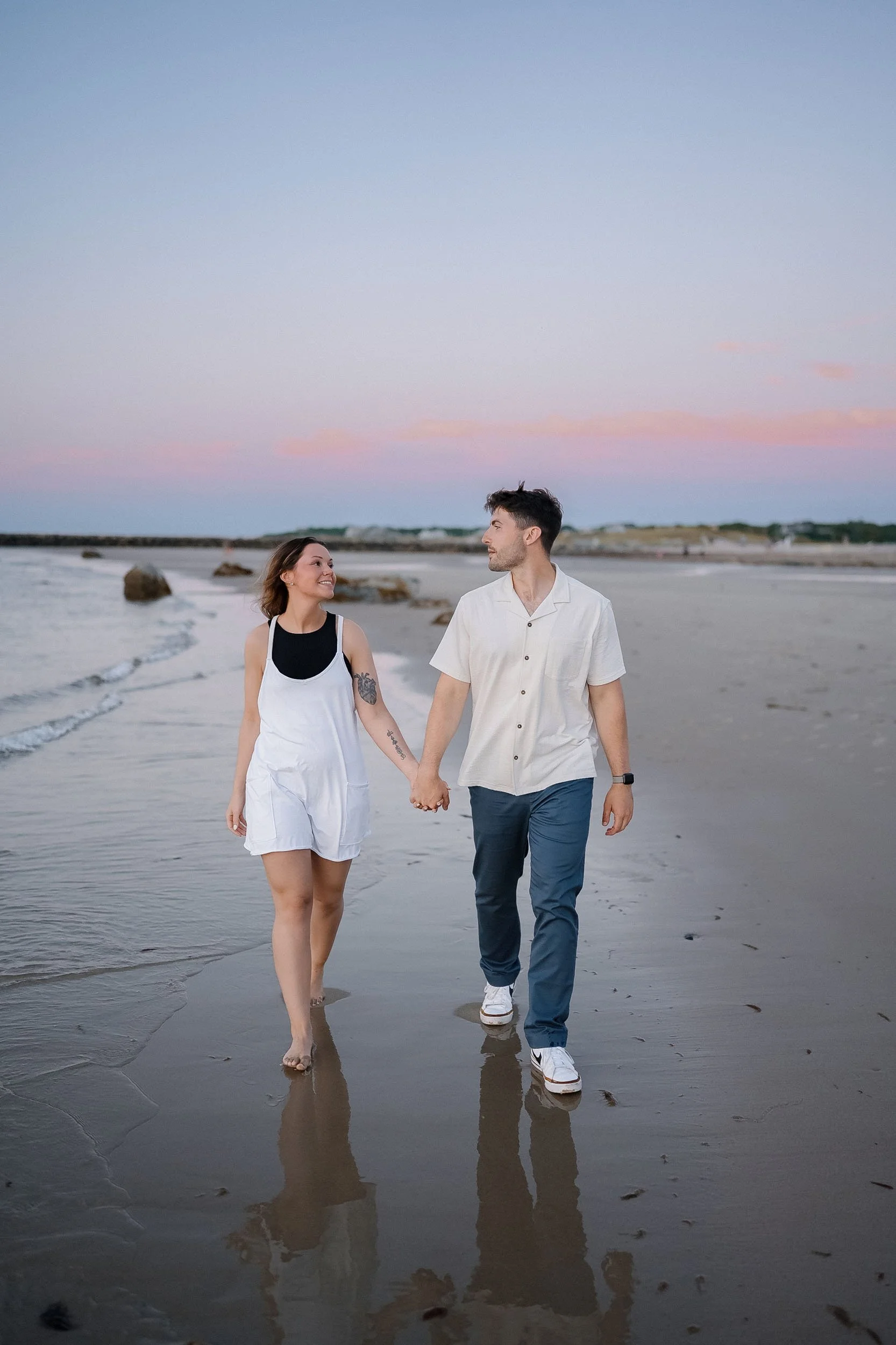 A couple holding hands and walking along the beach at sunset, with waves and rocks in the background.