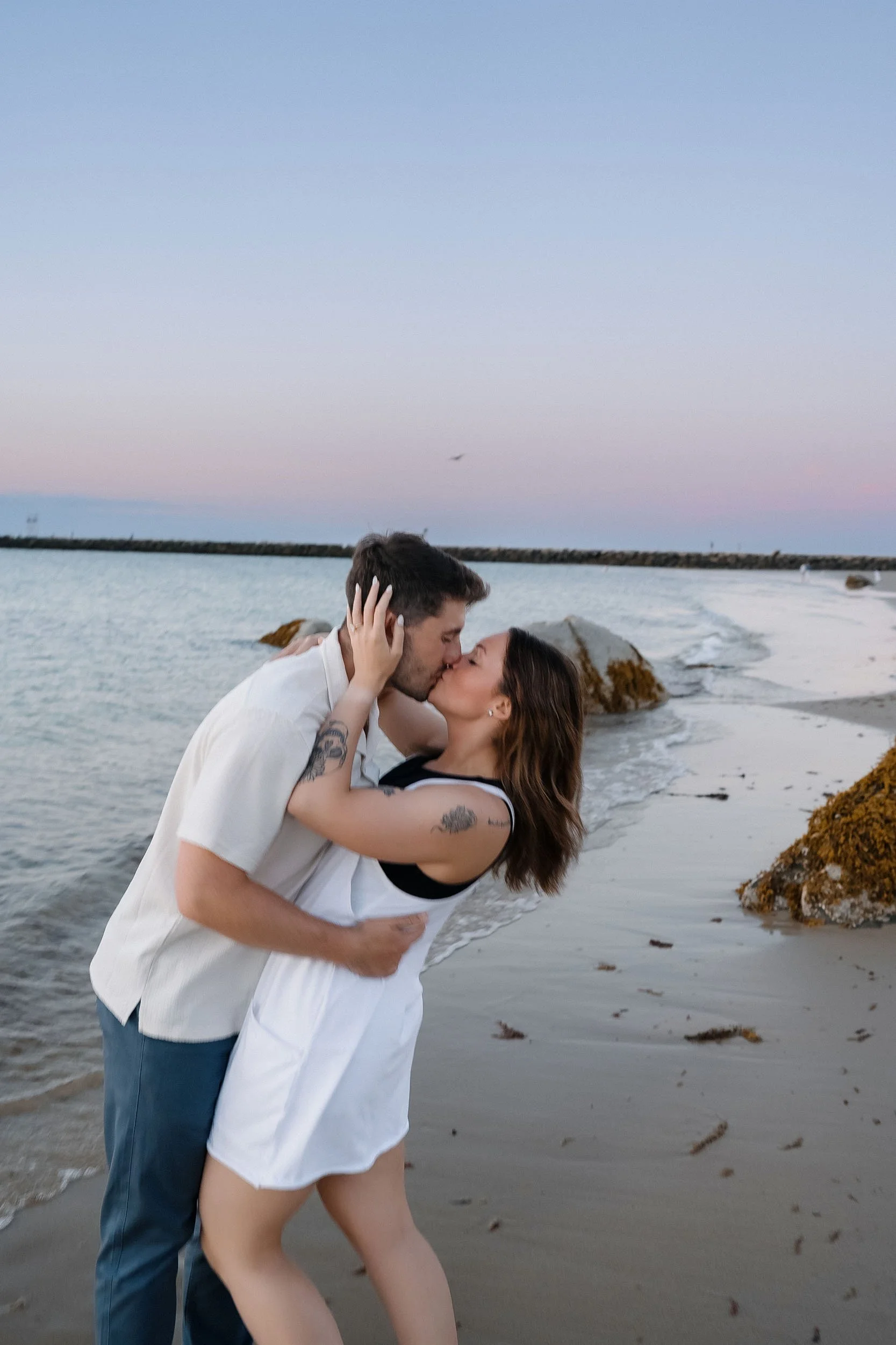 A couple kissing on the beach during sunset, with rocks and the ocean in the background.