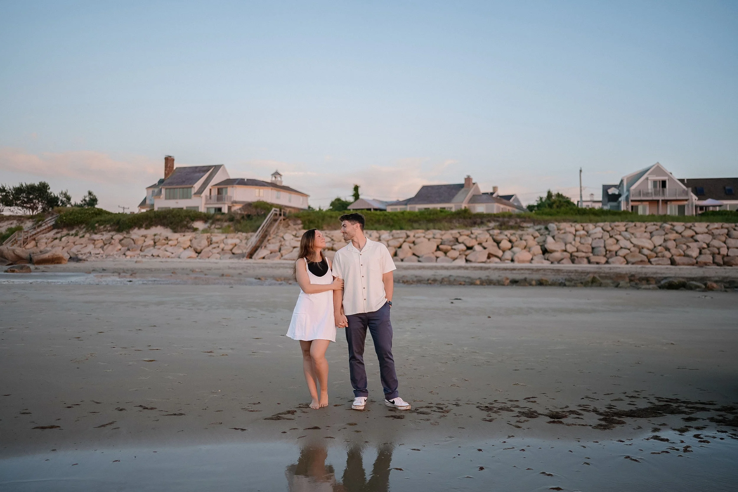 A couple holding hands on a beach at sunset with houses in the background.