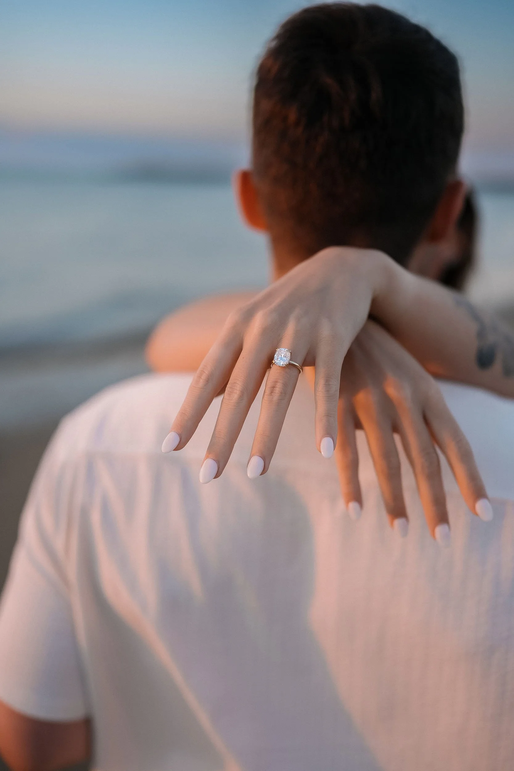 Close-up of a woman's hand with white nails, wearing a large diamond ring, resting on a man's shoulder at the beach during sunset.