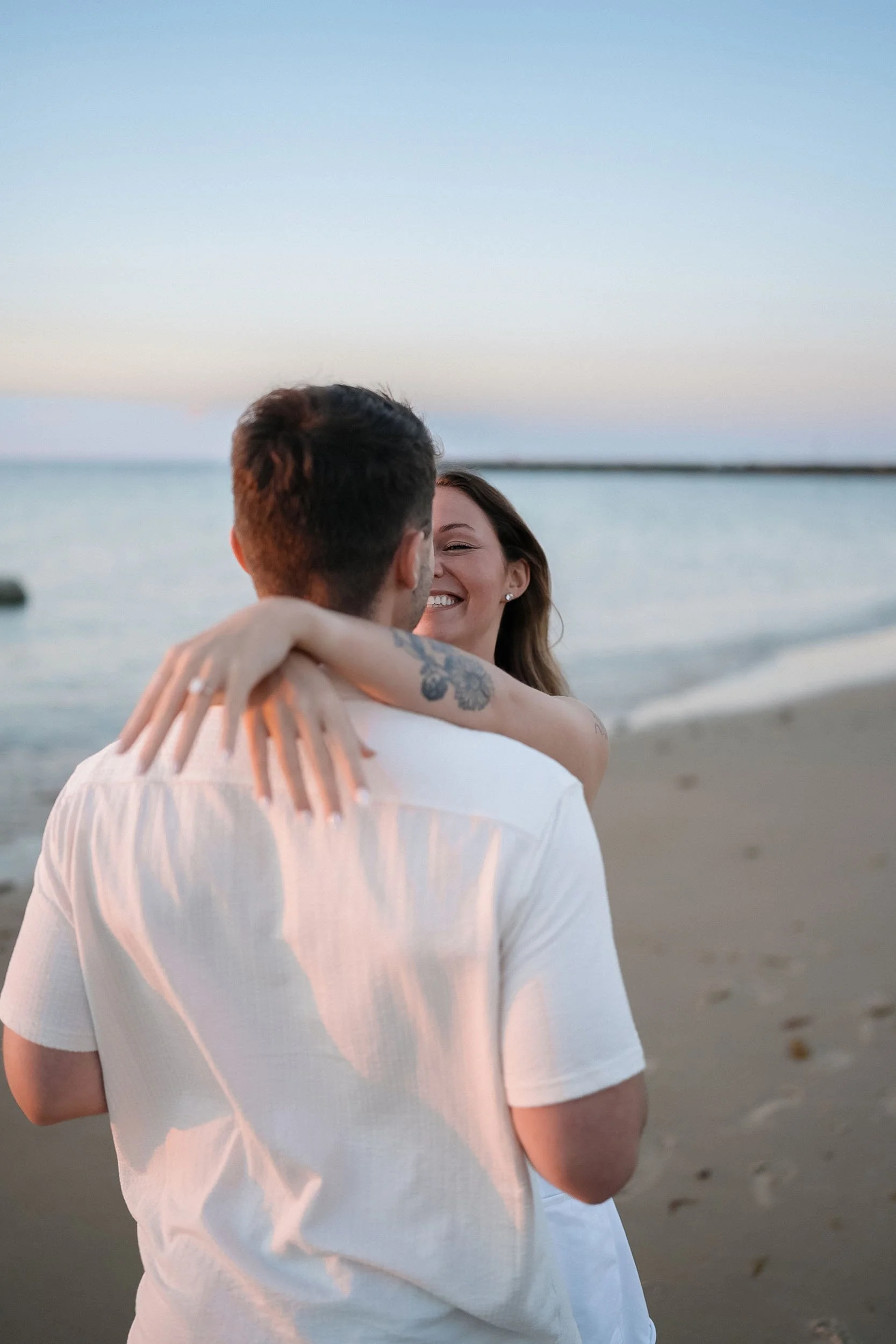 A joyful woman with long brown hair hugging a man with short dark hair on a beach at sunset, ocean in background.