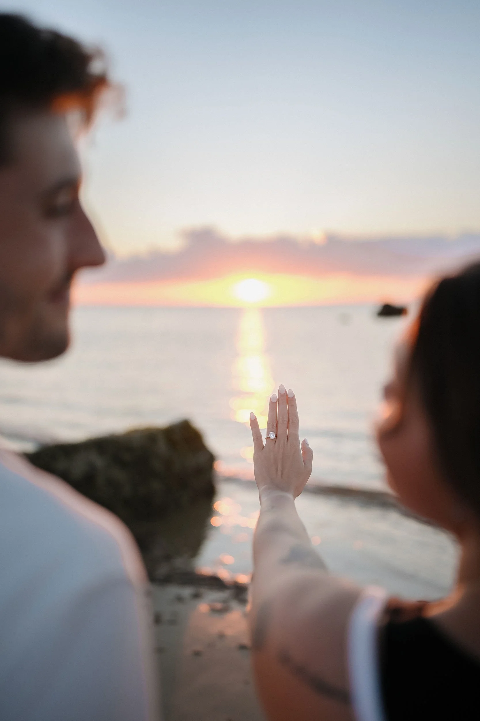 A couple on the beach during sunset, with the woman's left hand showing an engagement ring as she raises her fingers, with the ocean and rocks in the background.
