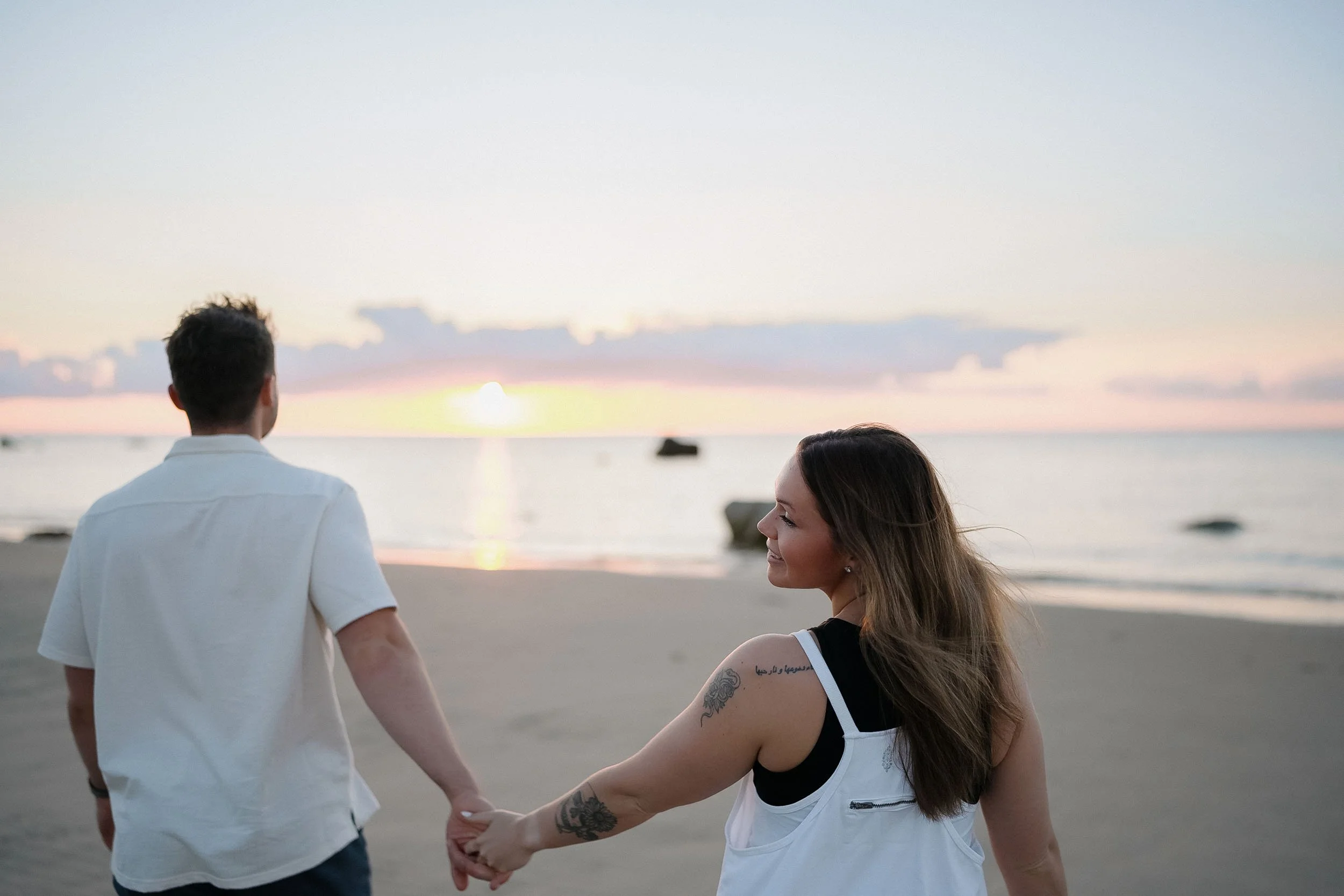 A young couple holding hands and walking on the beach during sunset, ocean in the background.