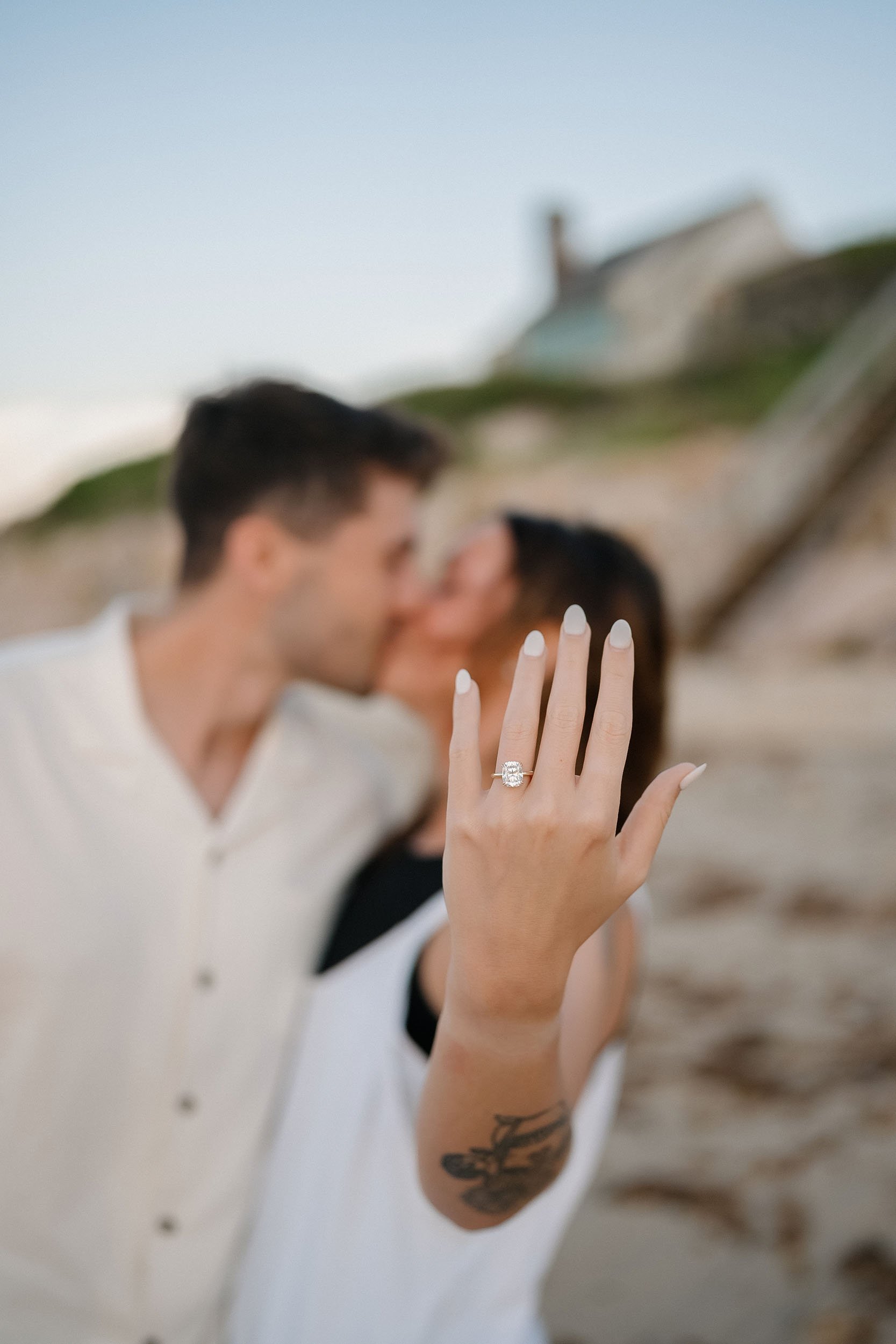 A woman showing off an engagement ring on her left hand with a blurred couple kissing in the background at the beach.