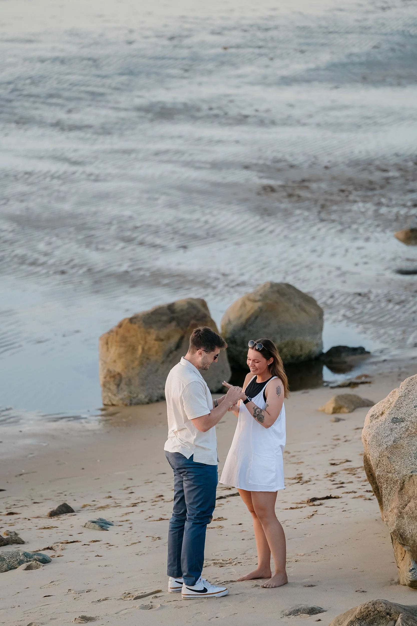 A couple standing on a sandy beach near large rocks, smiling and looking at a phone together during sunset.