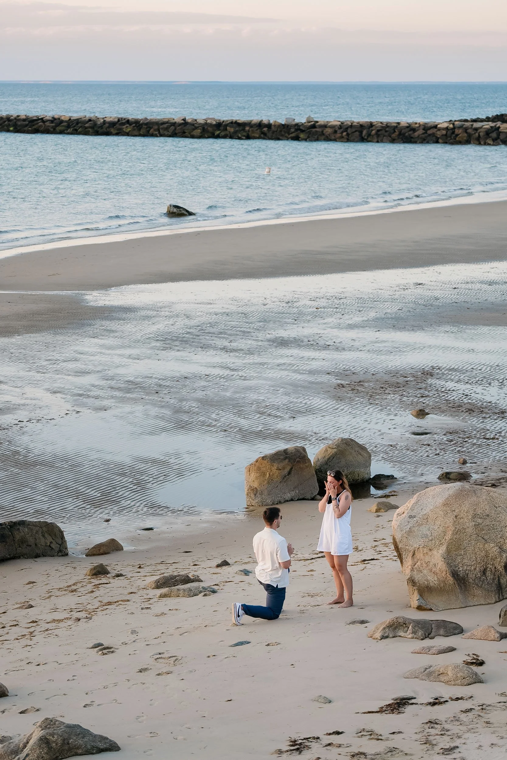 A man proposing marriage to a woman on a beach at sunset, with the ocean and rocks in the background.