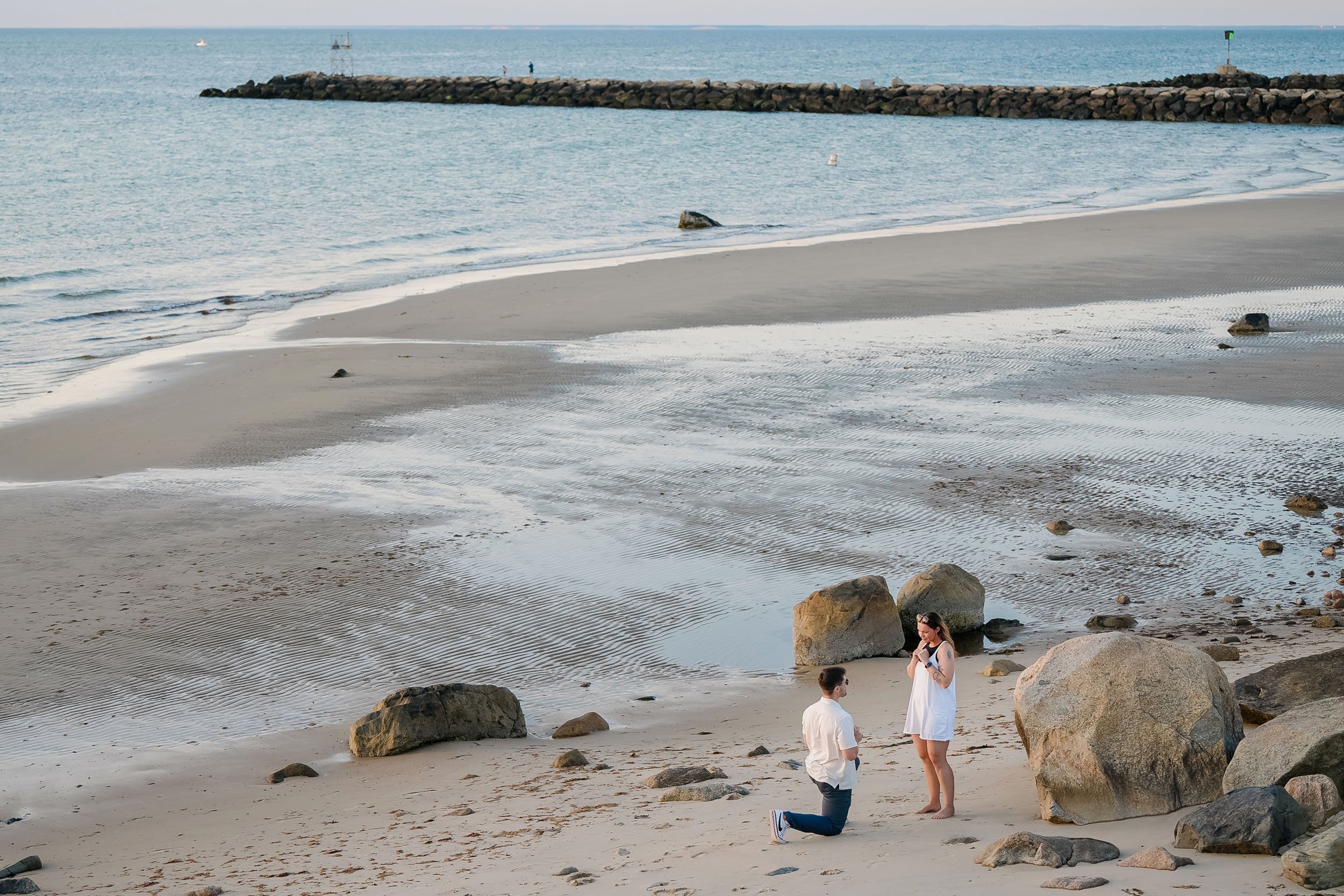 A man kneeling on one knee proposing marriage to a woman on a sandy beach with rocks, with the ocean and a pier in the background.