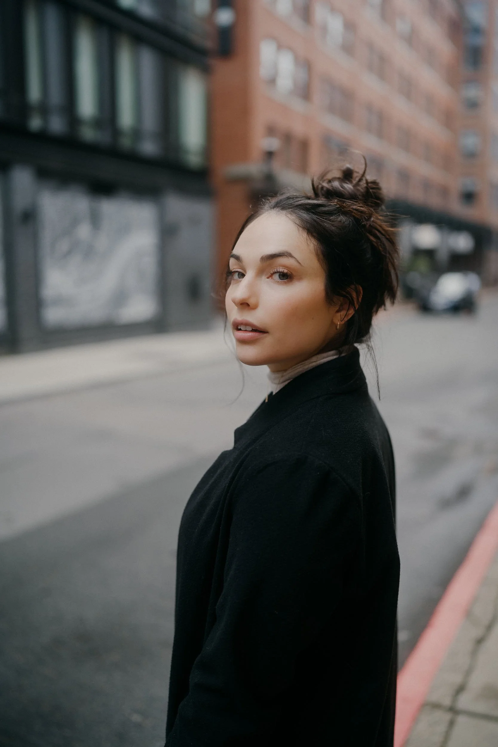 A young woman with dark hair styled in a messy bun, wearing a black coat and a beige turtleneck, standing on an urban street with brick apartment buildings in the background.