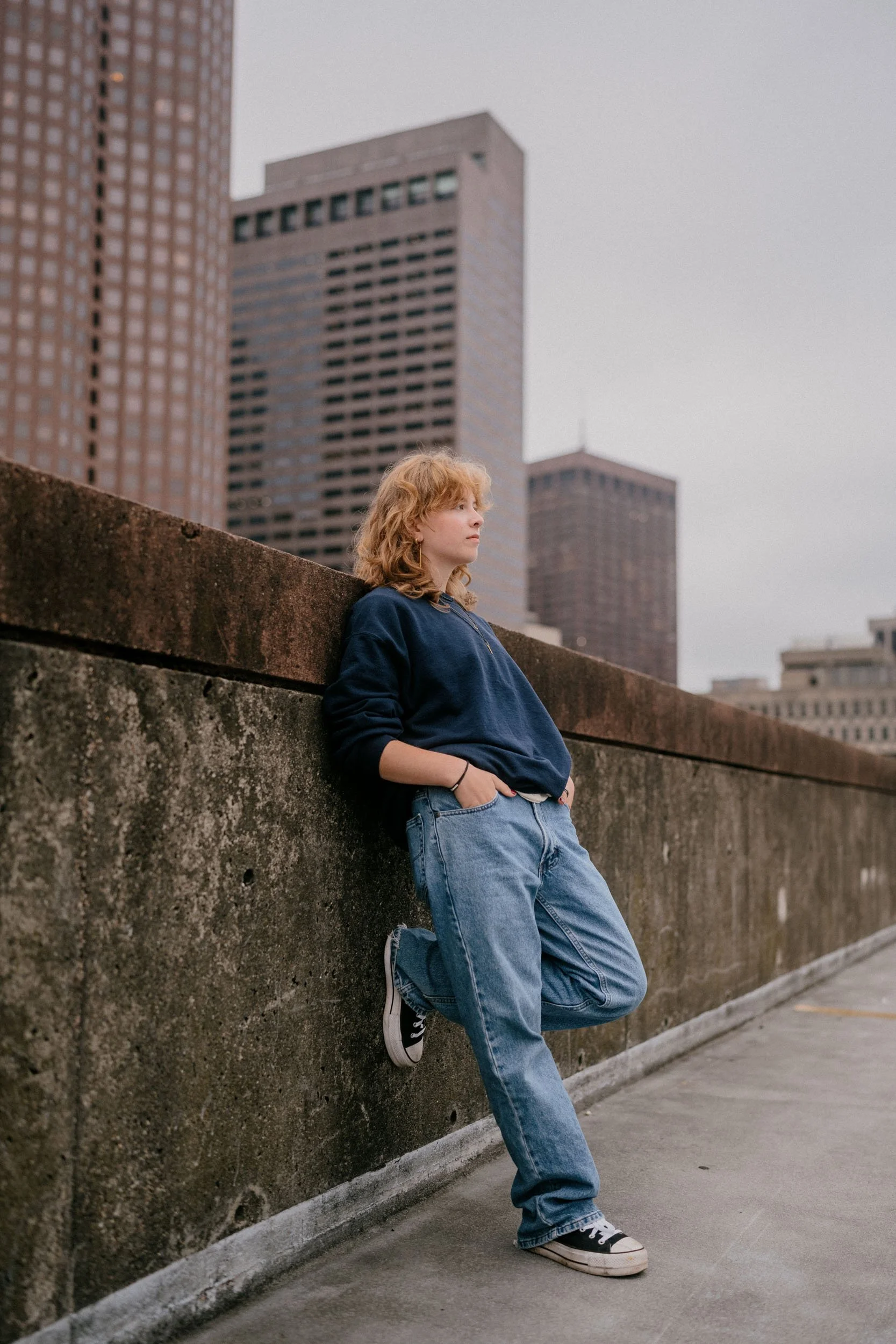 A young woman with curly red hair wearing a blue hoodie and jeans, leaning against a concrete wall on a city rooftop with skyscrapers in the background under a cloudy sky.
