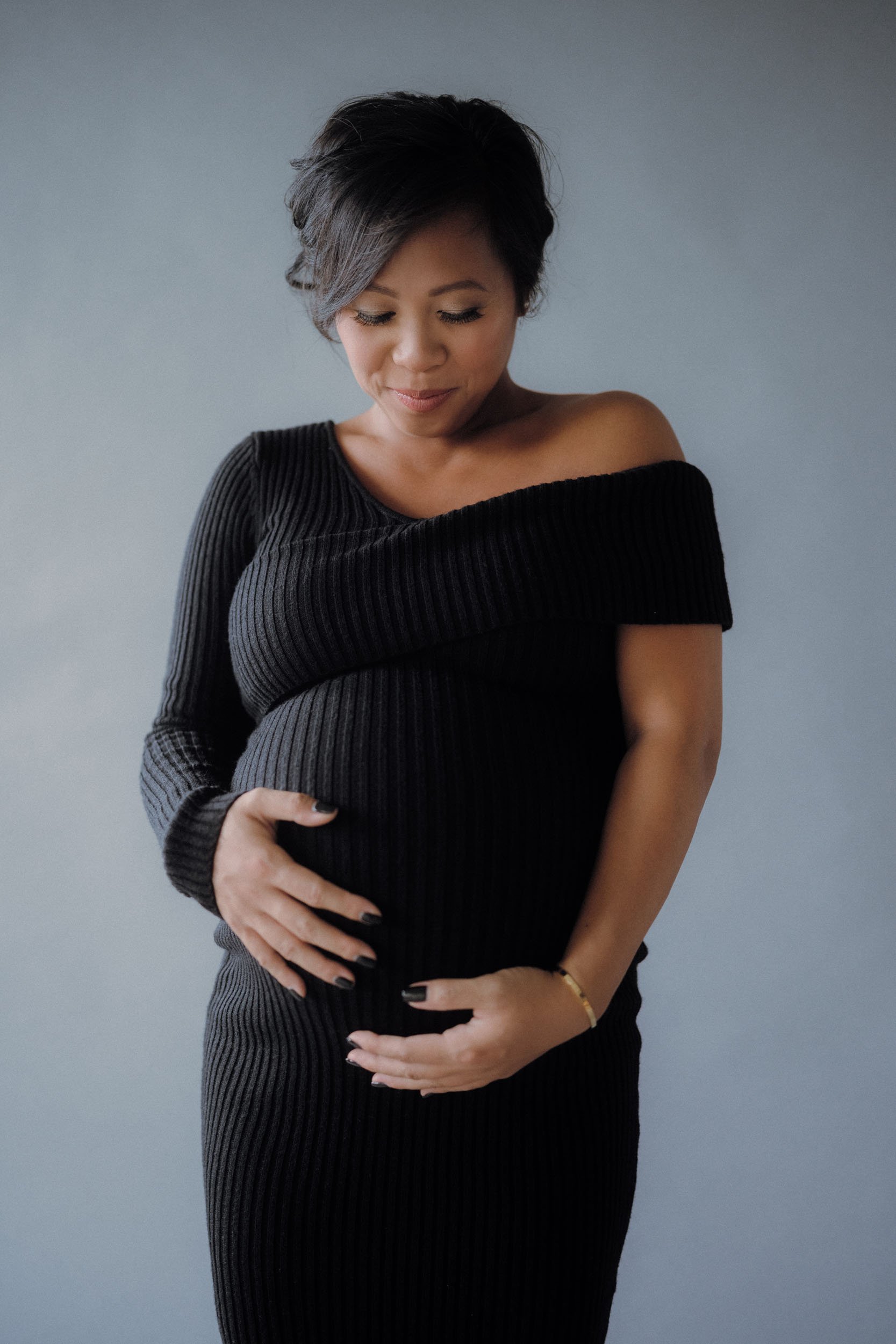 A pregnant woman wearing a black ribbed off-the-shoulder dress, standing against a plain gray background, looking down with hands on her belly.
