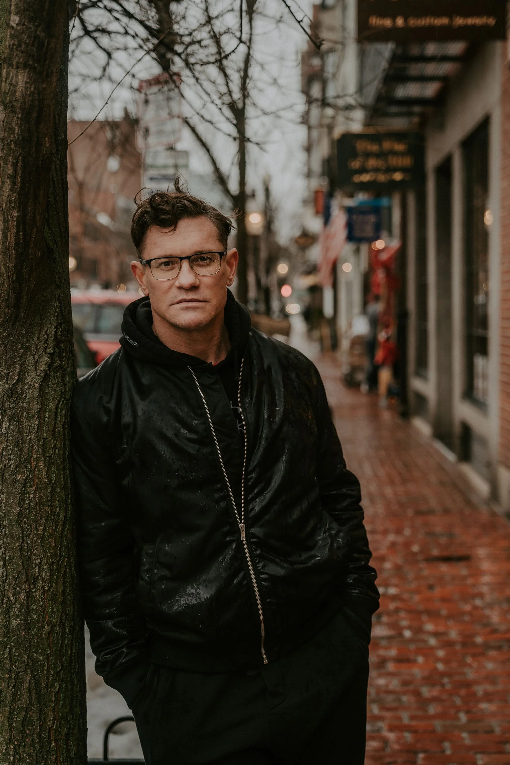 A man with glasses and dark hair stands leaning against a tree on a wet city sidewalk in the rain, wearing a black shiny jacket and black pants.