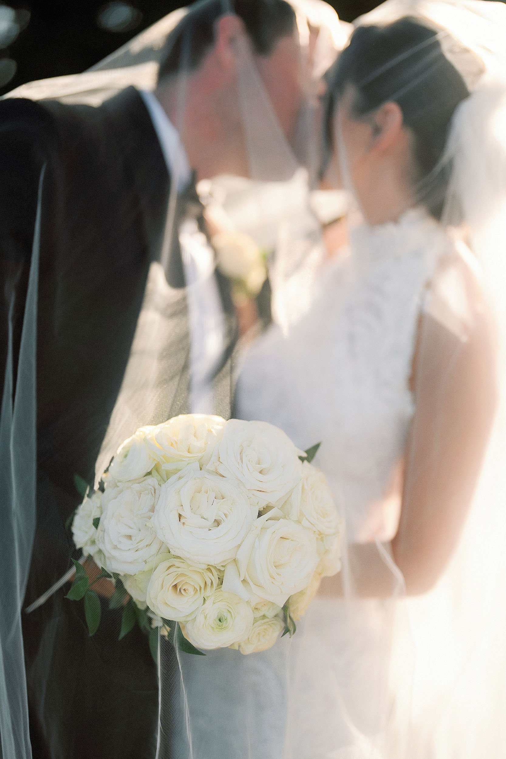A bride and groom lean in for a kiss, holding a bouquet of white roses, with sunlight creating a warm glow around them.