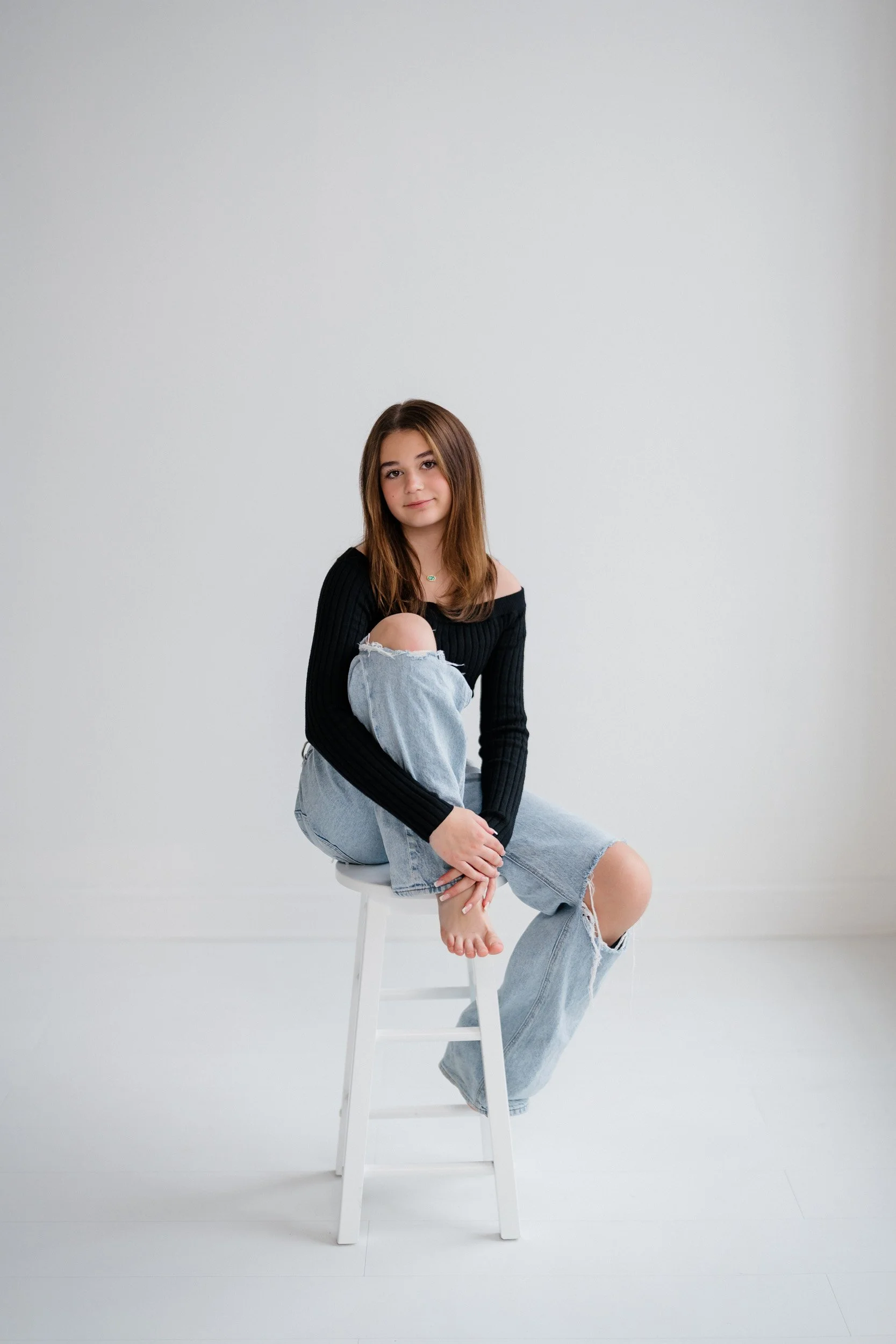 A young woman with long brown hair sitting on a white stool in a minimal white room, wearing a black off-shoulder sweater and ripped jeans, looking at the camera with a slight smile.