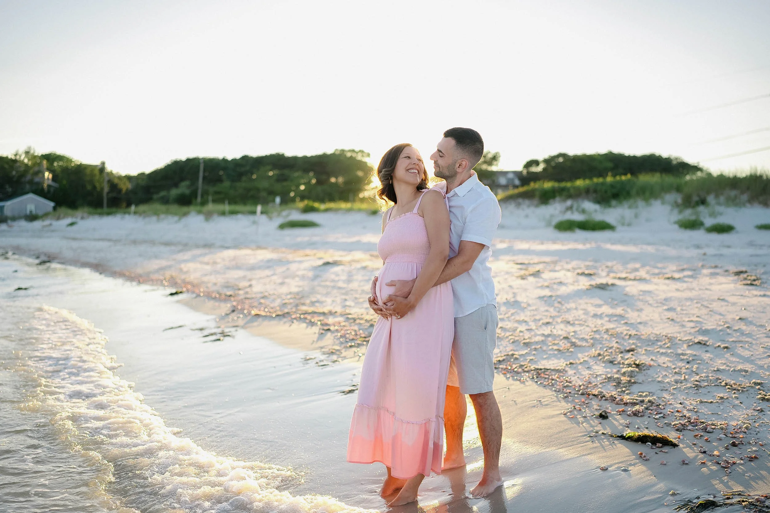 A couple stands on the beach at sunset, embracing and smiling at each other, with trees and houses in the background.