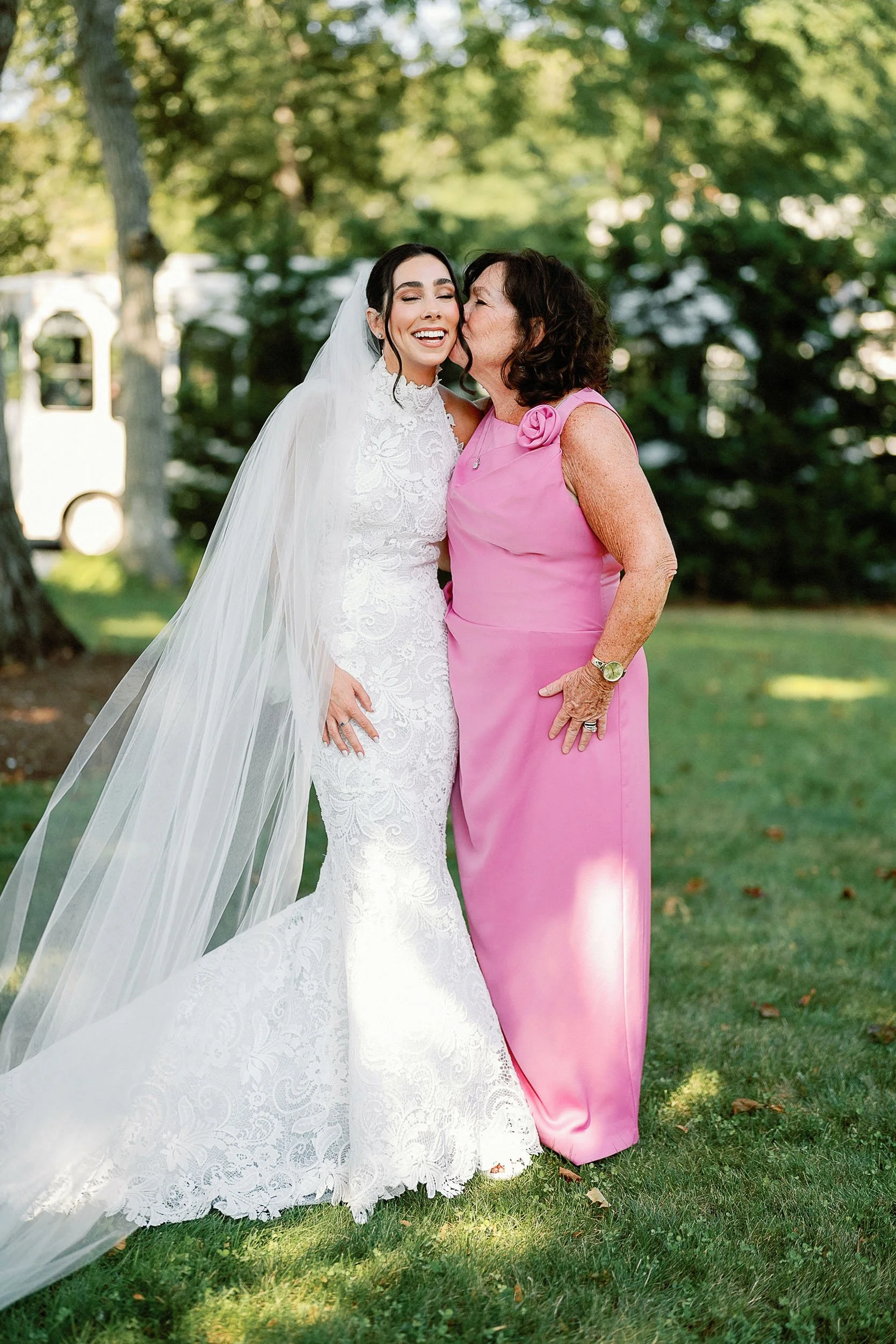 A bride in a white lace wedding dress and veil kisses an older woman in a pink dress on a sunny outdoor lawn surrounded by trees.