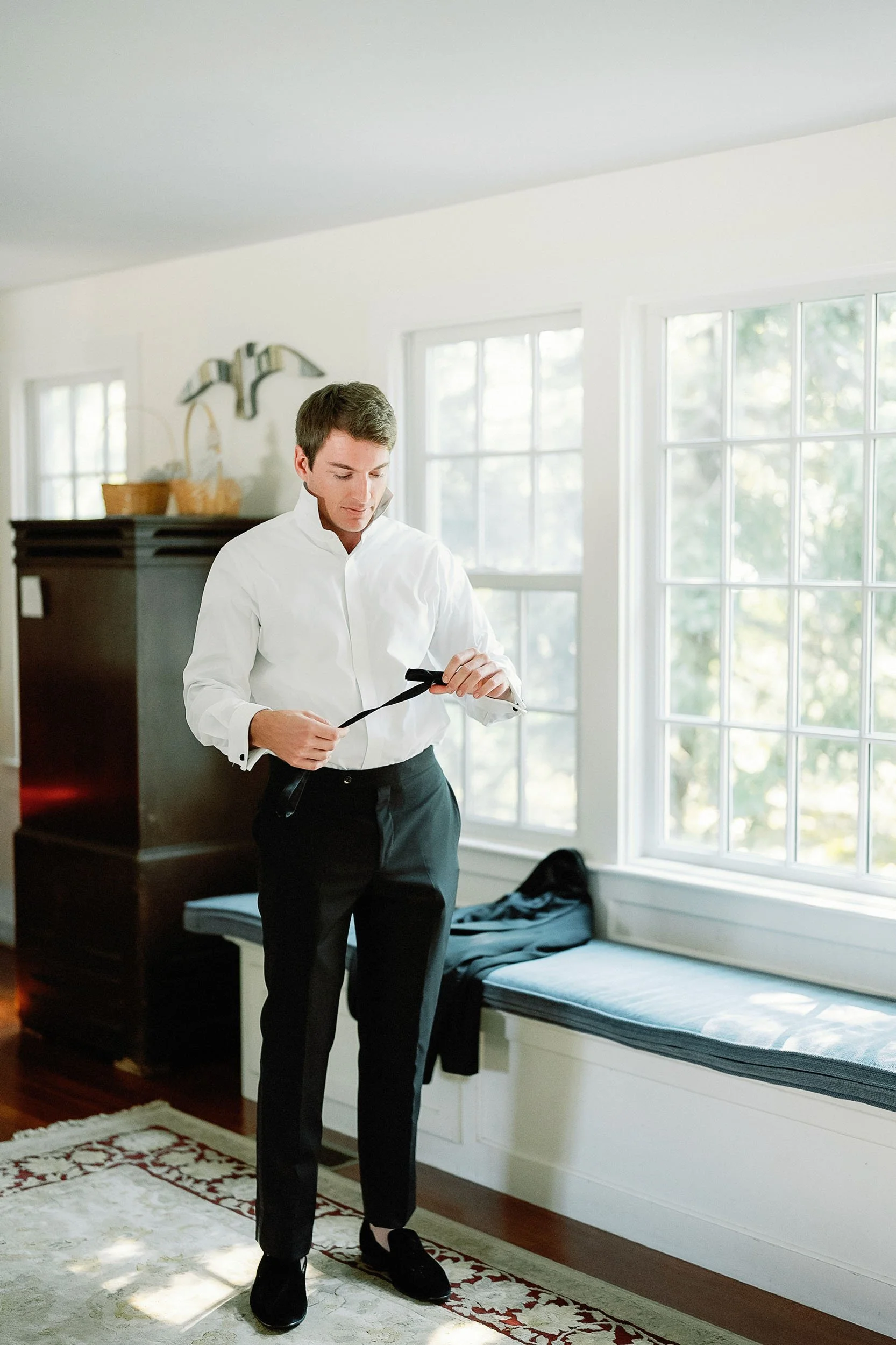 A man in a white dress shirt and black pants is tying a black bow tie indoors near a large window, with a bench underneath and sunlight streaming in.
