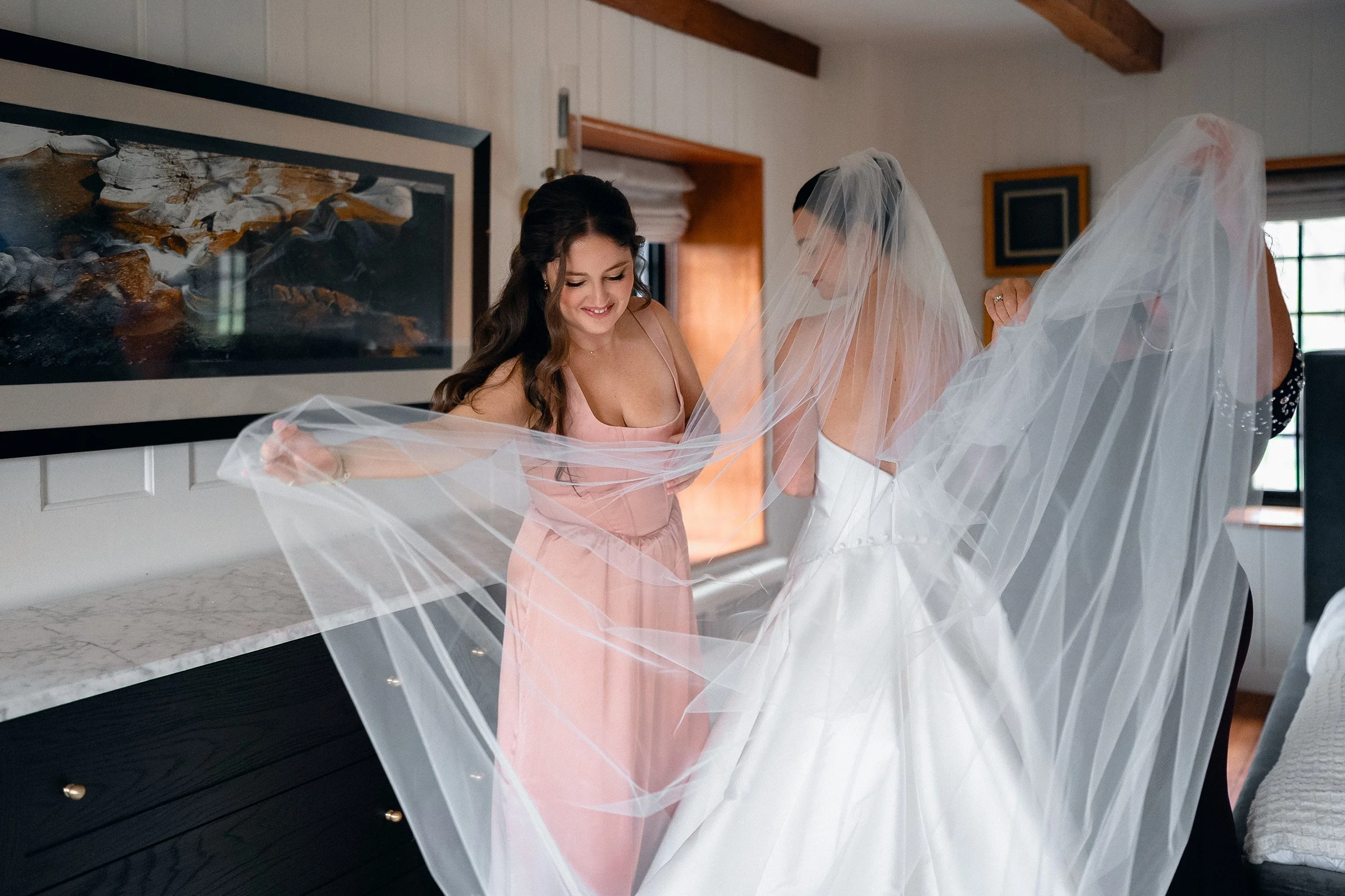 A bride in a white wedding dress with a veil standing with two women, one in a pink dress, holding the veil.