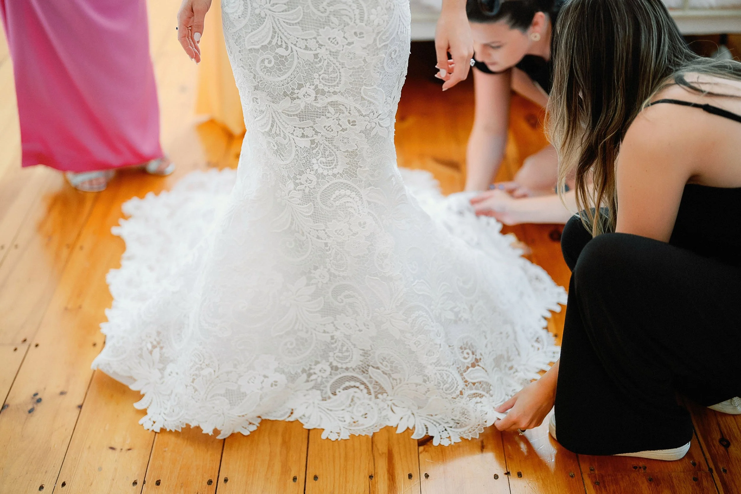 People helping a bride with the train of her white lace wedding gown on a wooden floor.