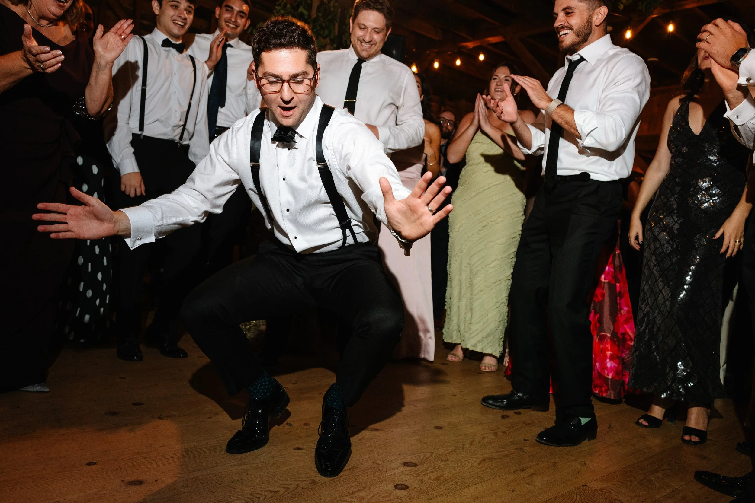A man in formal attire dancing energetically at a party surrounded by smiling guests.