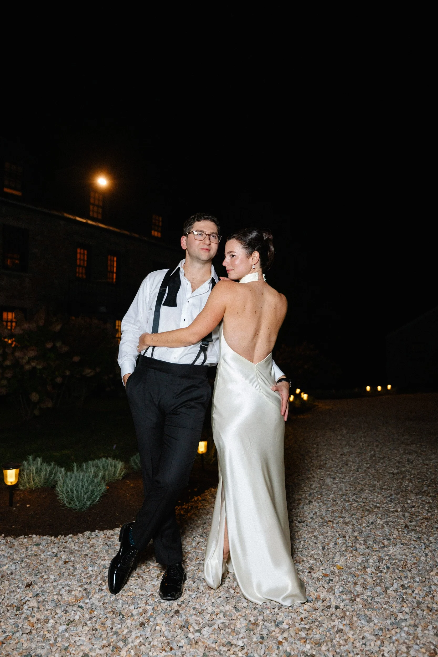 A couple dressed in formal attire poses outdoors at night, with the woman in a backless white satin gown and the man in a white shirt with suspenders, standing on a gravel path illuminated by ground lights.
