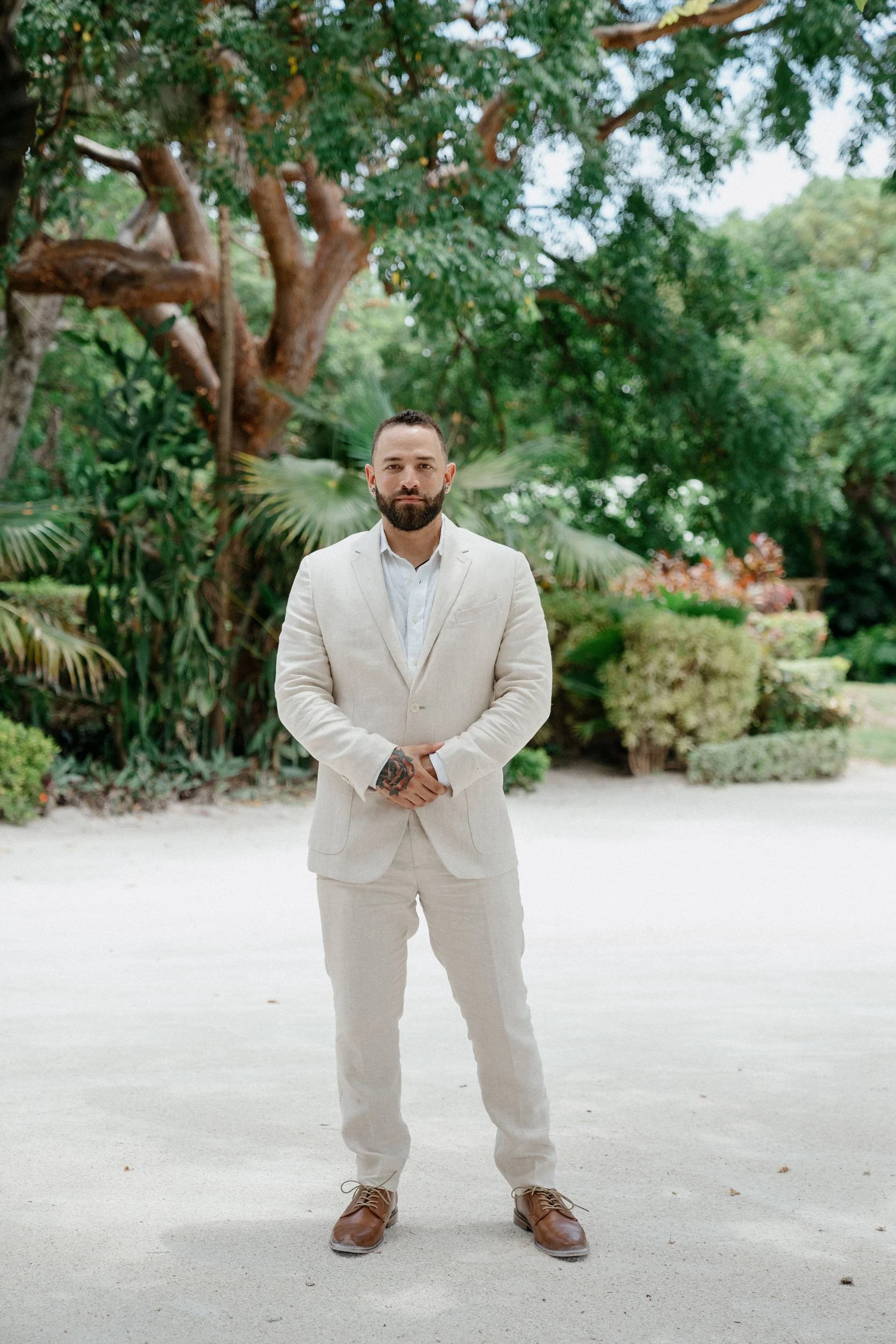 A man in a cream-colored suit with a white shirt, standing outdoors on a sandy path with lush green trees and bushes in the background.