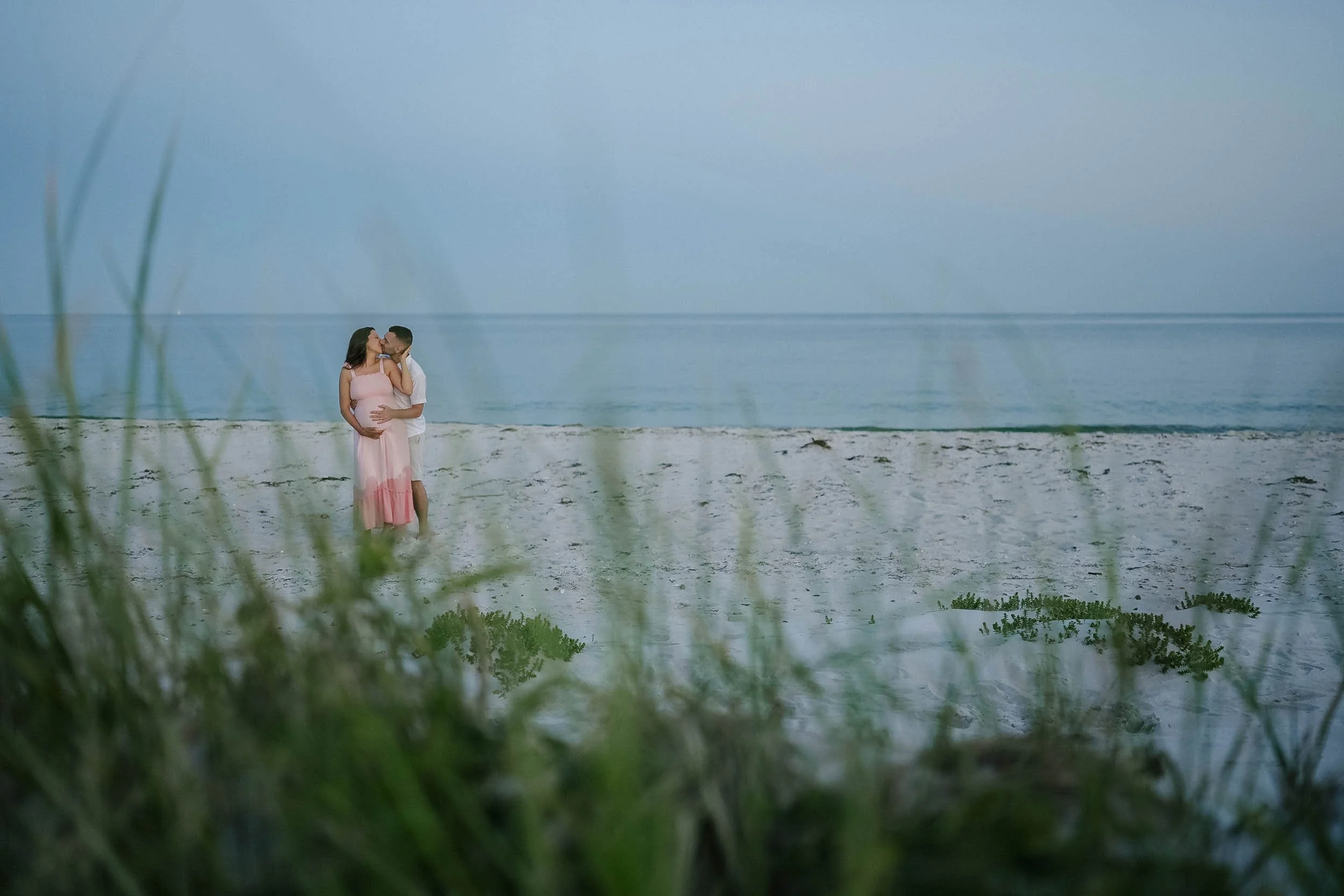 A couple in romantic embrace and kiss on a quiet sandy beach with grasses in the foreground, ocean in the background, and a soft blue sky.