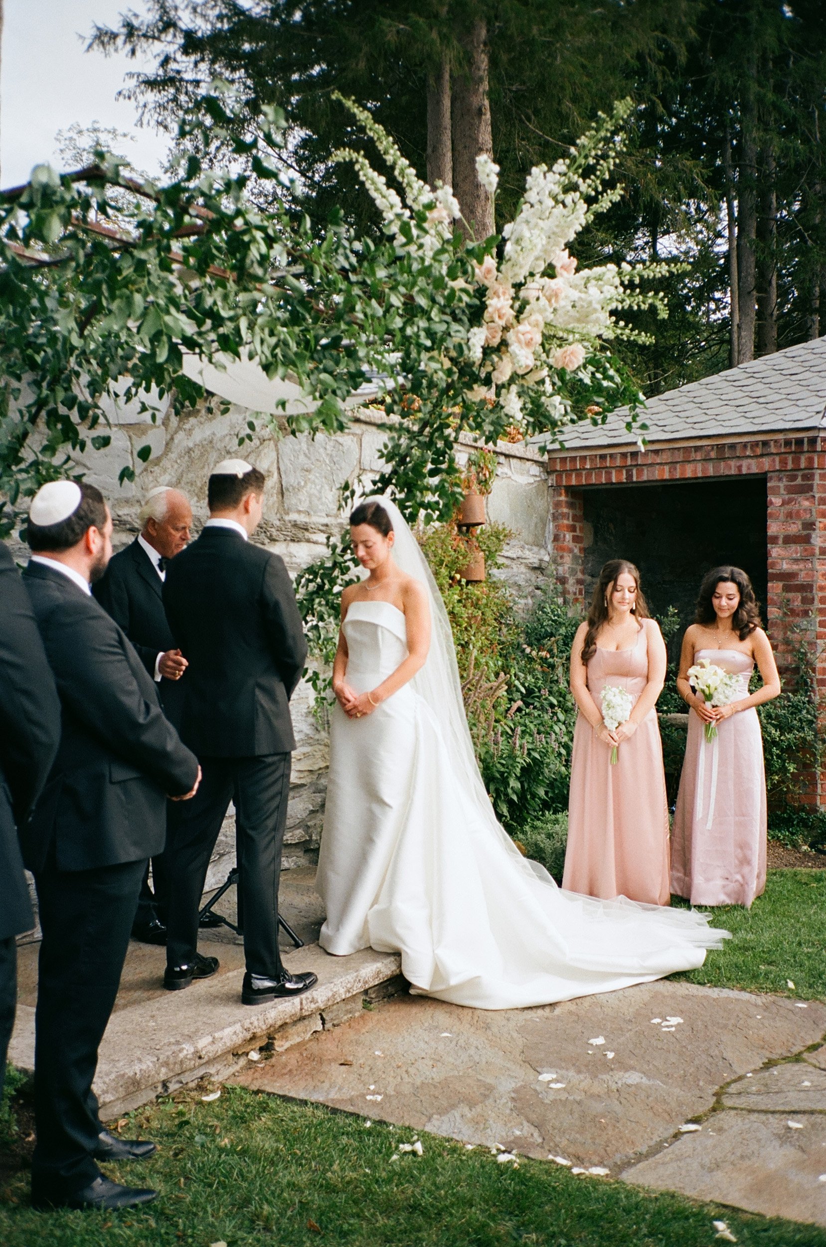 A wedding ceremony outdoors with the bride, groom, officiant, and two bridesmaids, under a floral arrangement of white and pale pink flowers and greenery.